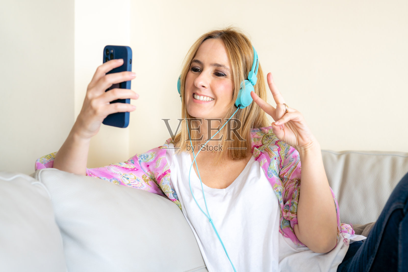 微笑的年轻女子戴着耳机坐在家里的沙发上使用智能手机。照片摄影图片