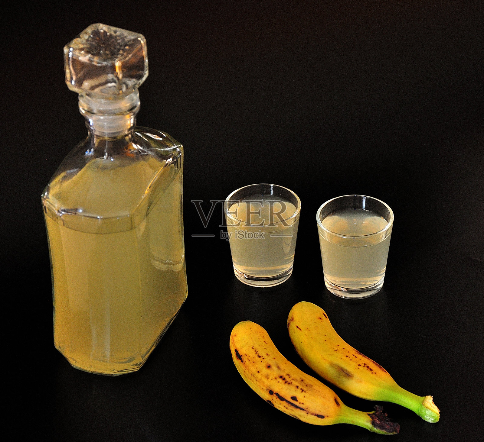 香蕉利口酒,一瓶自制烈酒和两个杯子在黑色的背景上,成熟的水果在附近。照片摄影图片