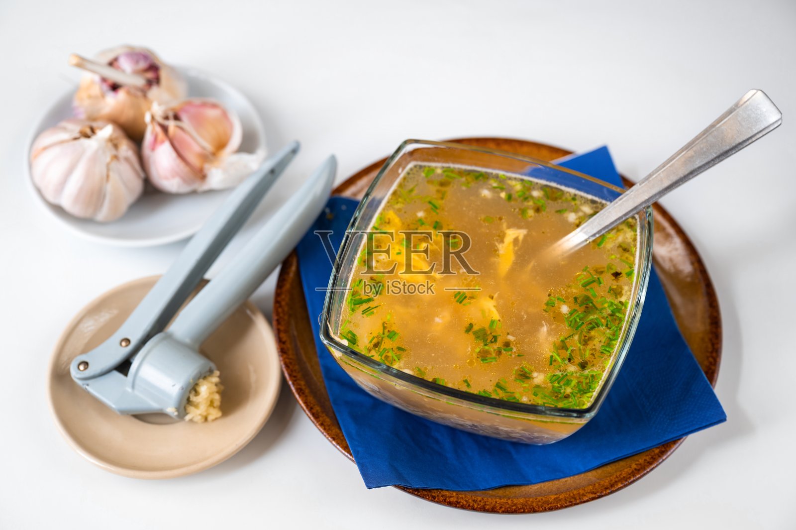 用玻璃碗盛蒜蛋肉汤,压蒜机和大蒜上桌。照片摄影图片