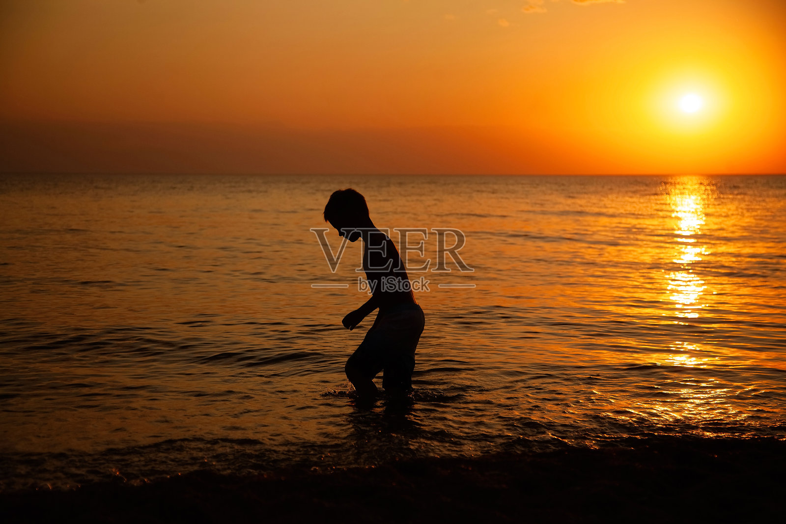 一个男孩站在海中,看着夕阳下的水里的东西,侧面的轮廓照片摄影图片