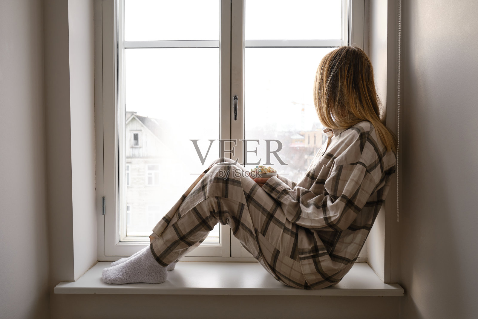 一个穿着睡衣的女孩坐在窗台上,手里拿着一杯加了棉花糖的可可。在家里度过舒适的冬日。检疫时间照片摄影图片