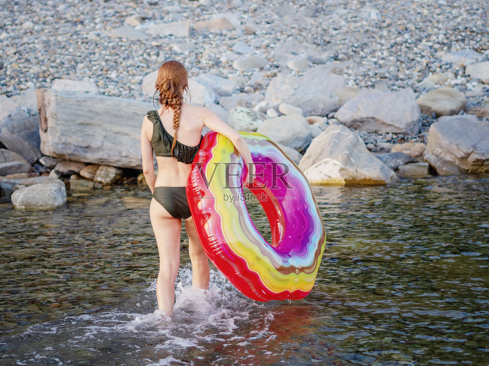 一名身穿湿式比基尼的女子手持一个巨大的彩色游泳圈,穿过海水走向海岸照片摄影图片