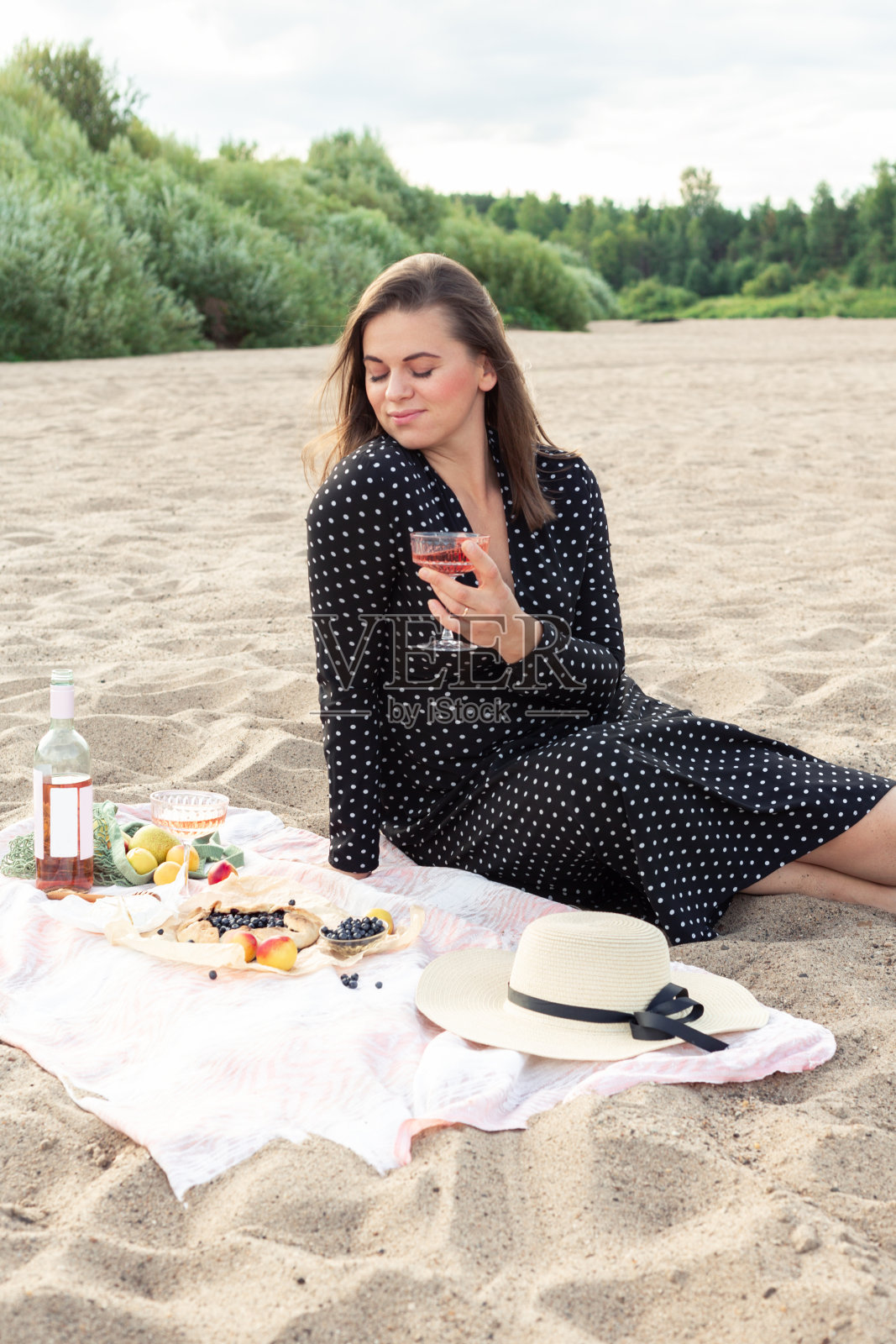 迷人的年轻女子拿着一杯玫瑰葡萄酒在沙滩上。玫瑰葡萄酒,各种水果,面包和奶酪照片摄影图片