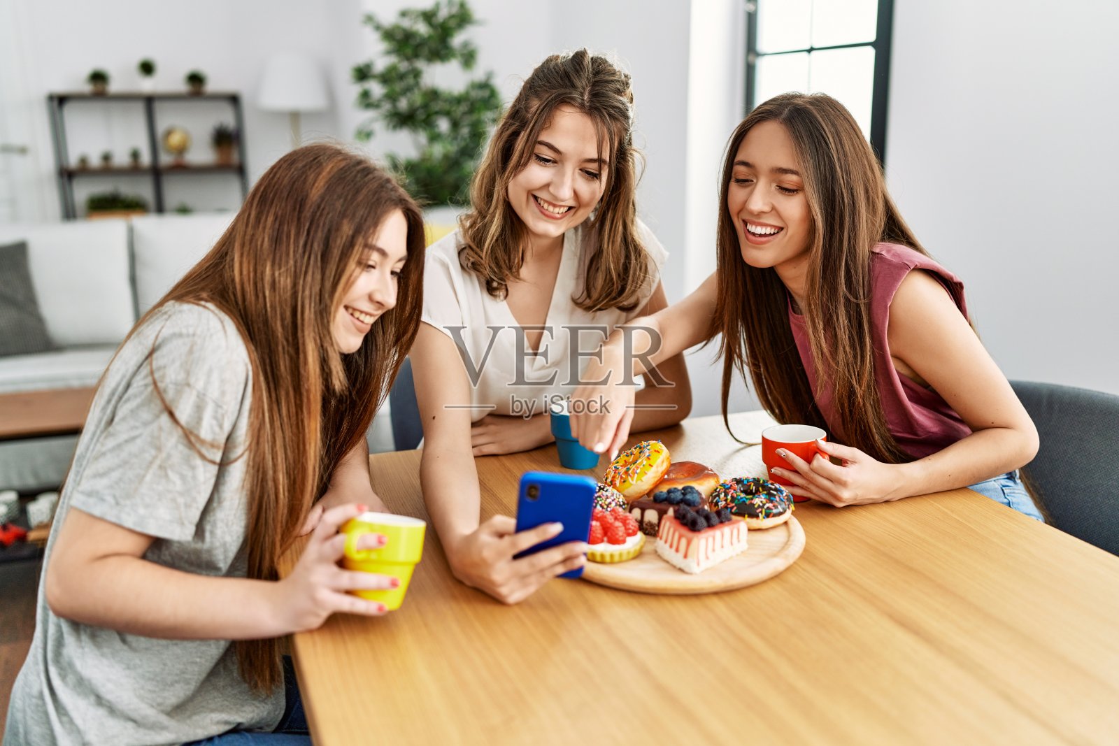 三个年轻的朋友坐在家里的桌子上,用智能手机吃早餐,笑得很开心。照片摄影图片
