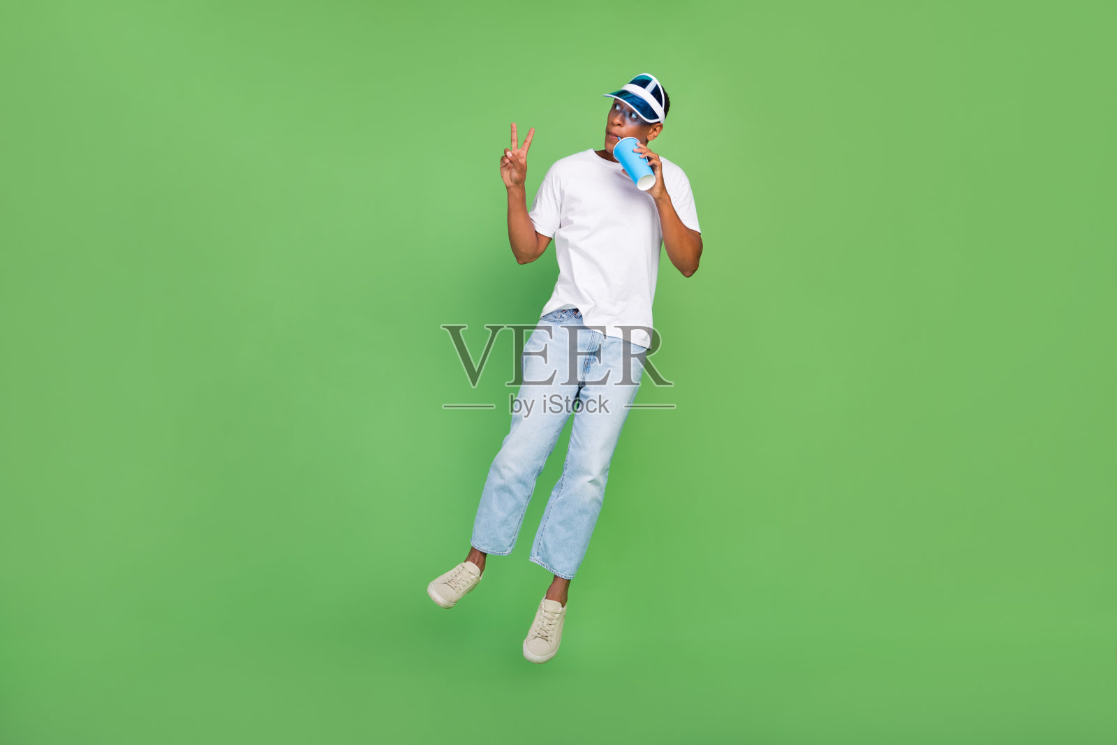 全尺寸照片的酷千禧一代深色的家伙喝饮料跳穿白色t恤牛仔裤鞋子孤立在绿色的背景照片摄影图片