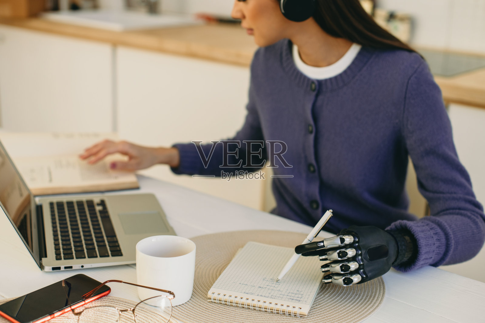 裁剪拍摄的忙碌的女学生在veri peri温暖的开襟羊毛衫坐在桌旁笔记本电脑和智能手机旁的拷贝空间屏幕,在仿生假肢手握笔,阅读书籍,做笔记照片摄影图片