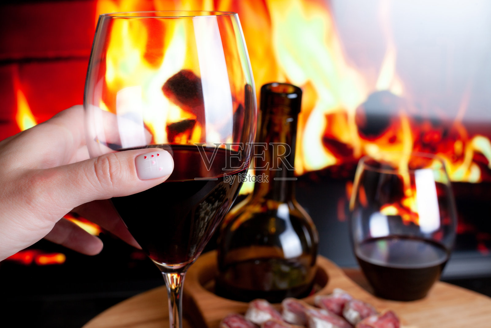 女性手握酒杯,靠在酒桌上,为葡萄酒和电壁炉准备开胃菜。用红心修指甲。浪漫的家庭晚餐概念照片摄影图片