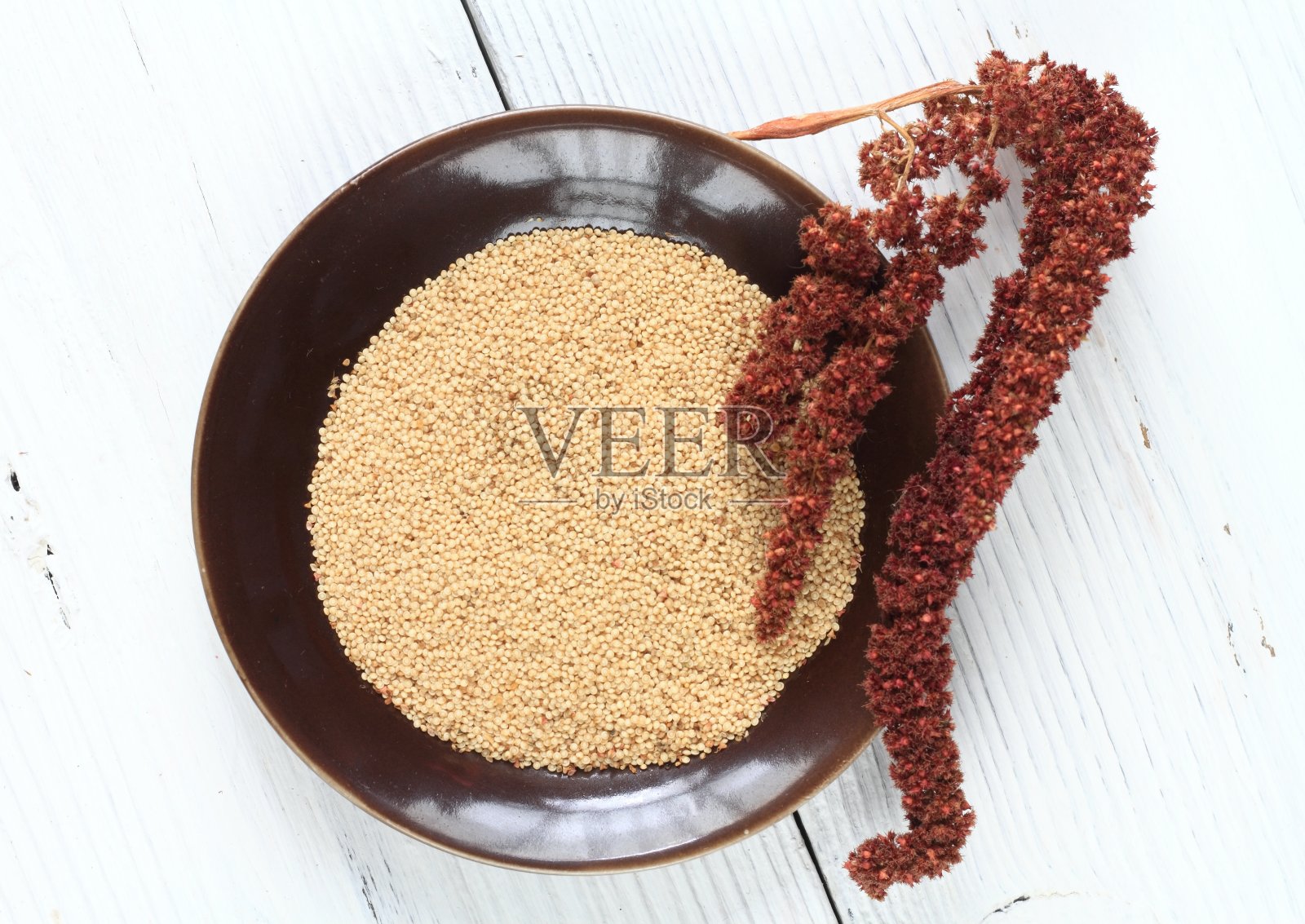 棕色盘子上放着苋菜种子,白色木桌上放着干红花。照片摄影图片