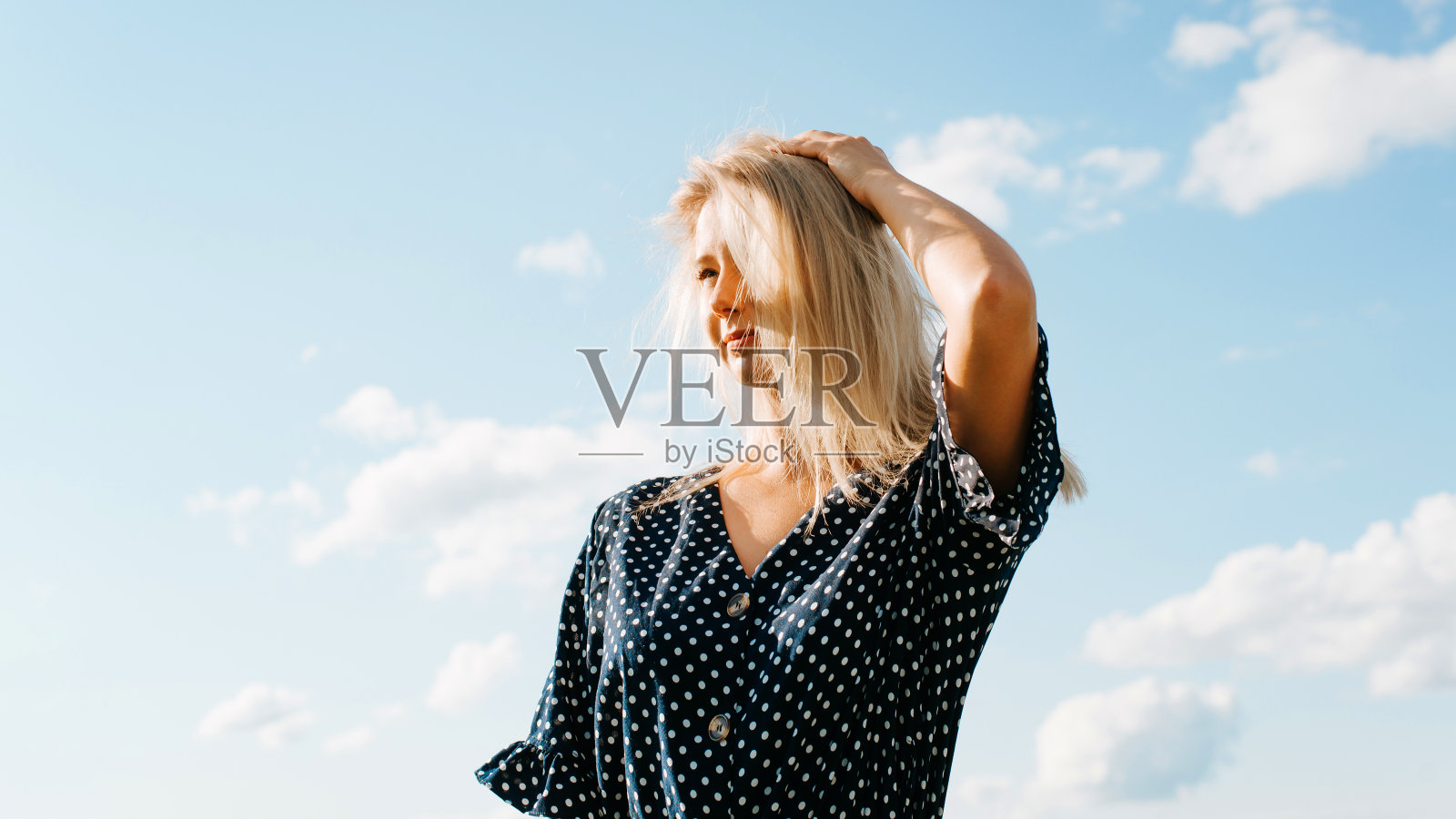 漂亮的年轻金发女人在波尔卡圆点夏季连衣裙反对蓝色多云的天空看向户外。漂亮的白人女孩的肖像模型在阳光明媚的大风天照片摄影图片