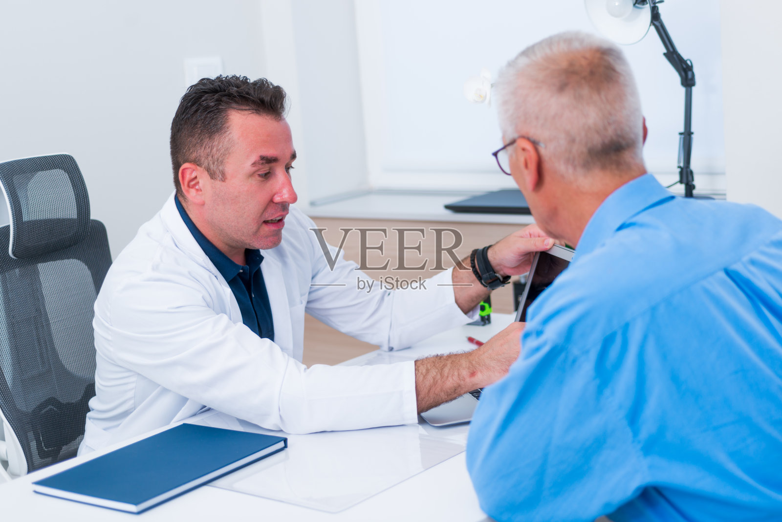 图为,一位中年医生坐在办公室里,用笔记本电脑向比他年长的病人展示自己的血检结果。照片摄影图片