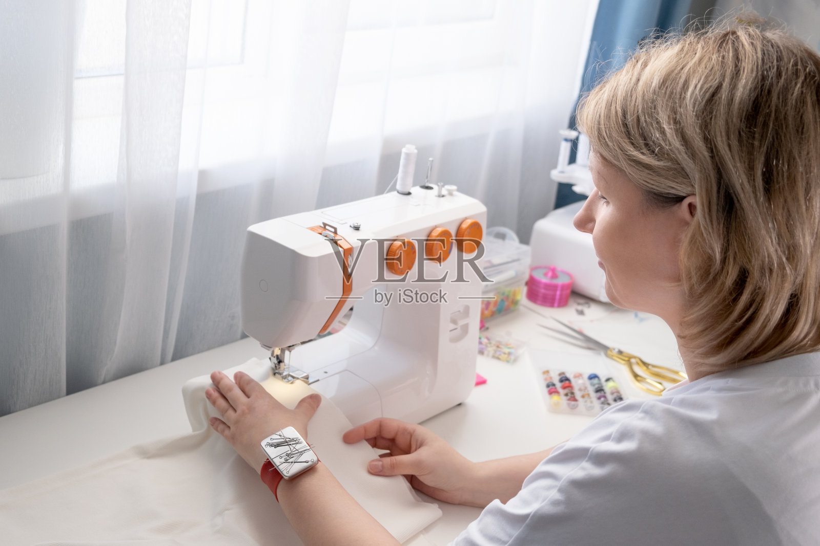 真实的中年妇女在家里用缝纫机缝制衣服。女裁缝师工作。在室内手工制作衣服。女性坐在那里享受缝制衣服的过程。创意技能设计,爱好和小型企业概念照片摄影图片