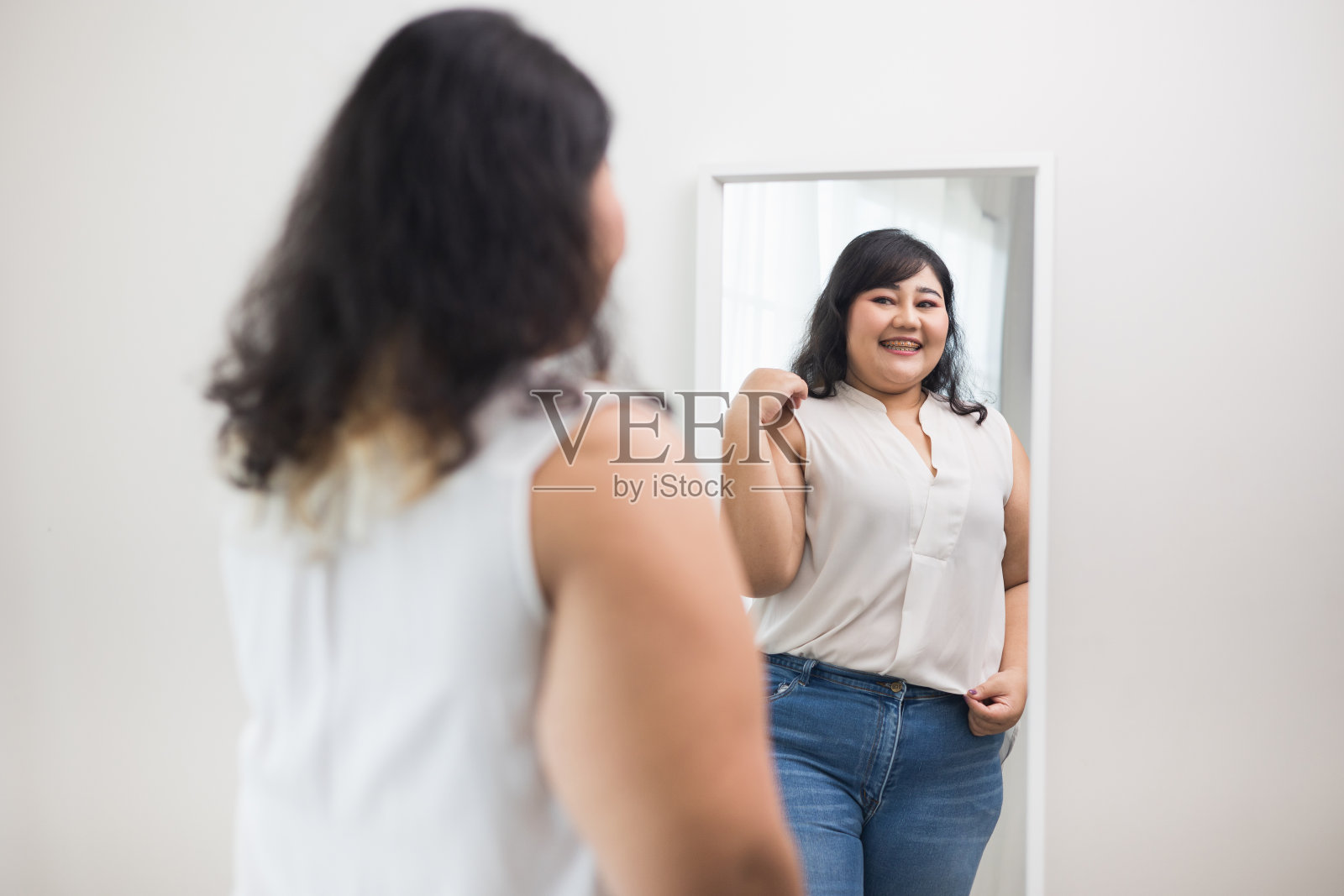 身体正面加尺码的女性服装。身材正胖的女人开心地看着家里的镜子,为自己感到骄傲。试着穿合身的衣服。照片摄影图片