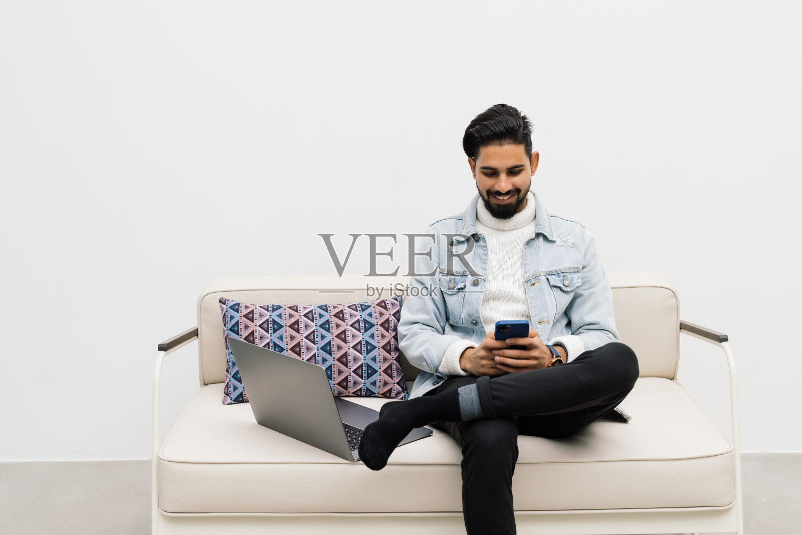 英俊的印度年轻男子坐在家里,手里拿着智能手机,和朋友聊天,在沙发上用手机放松,照片摄影图片