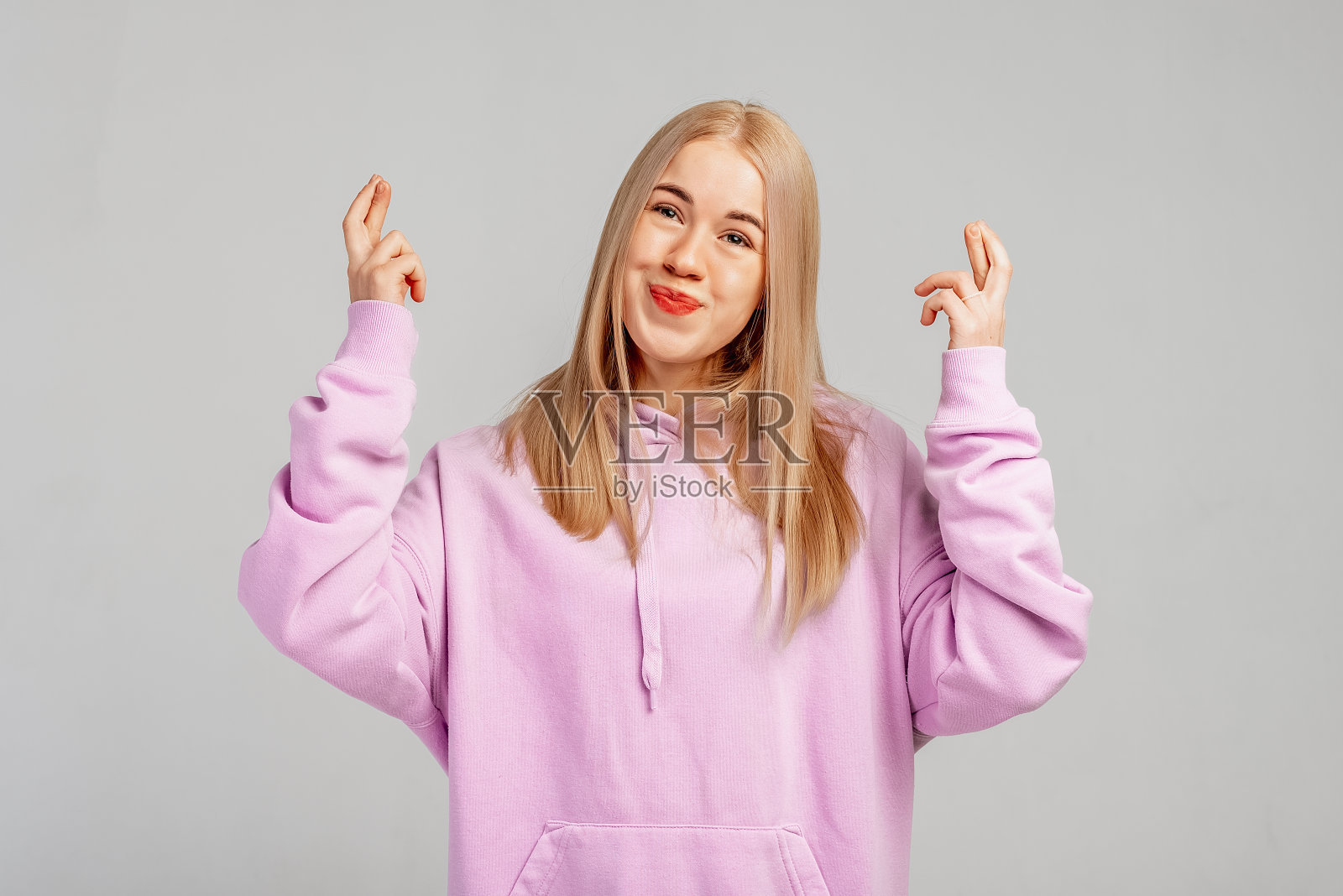 年轻的金发女子穿着紫色连帽衫,背景灰色,手指交叉,微笑着希望,梦想成真的信念。运气与迷信观念照片摄影图片