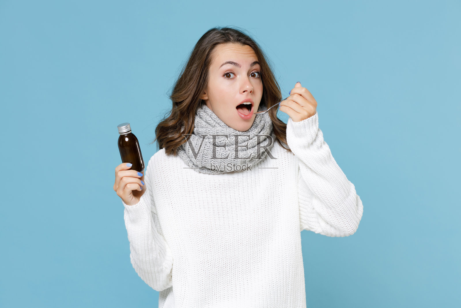 震惊的年轻女子穿着白色毛衣灰色围巾拿勺子液体药进瓶子孤立在蓝色背景工作室肖像。健康的生活方式治疗寒季病的观念。照片摄影图片