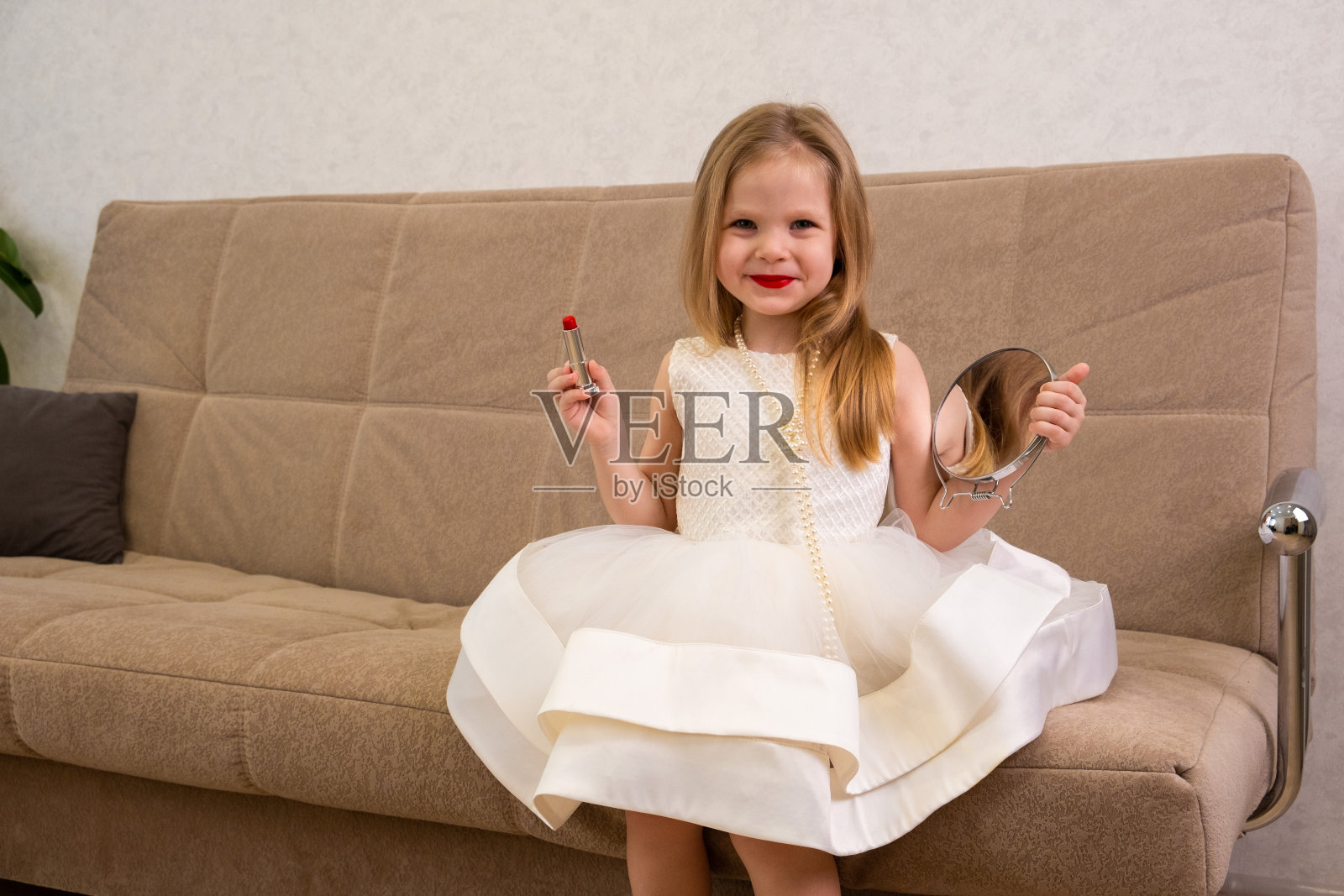 一个涂着红色嘴唇的金发小女孩,穿着白色的裙子、珠子和鞋子,坐在房间的沙发上,拿着口红和镜子照片摄影图片