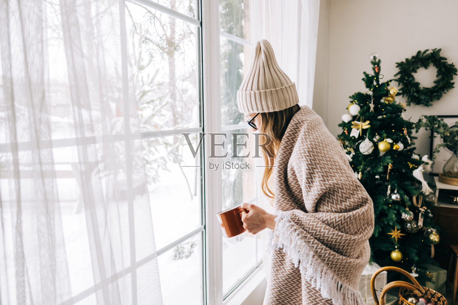 年轻的白人妇女站在窗边,看着窗外,喝着茶,享受着冬天的时光。照片摄影图片
