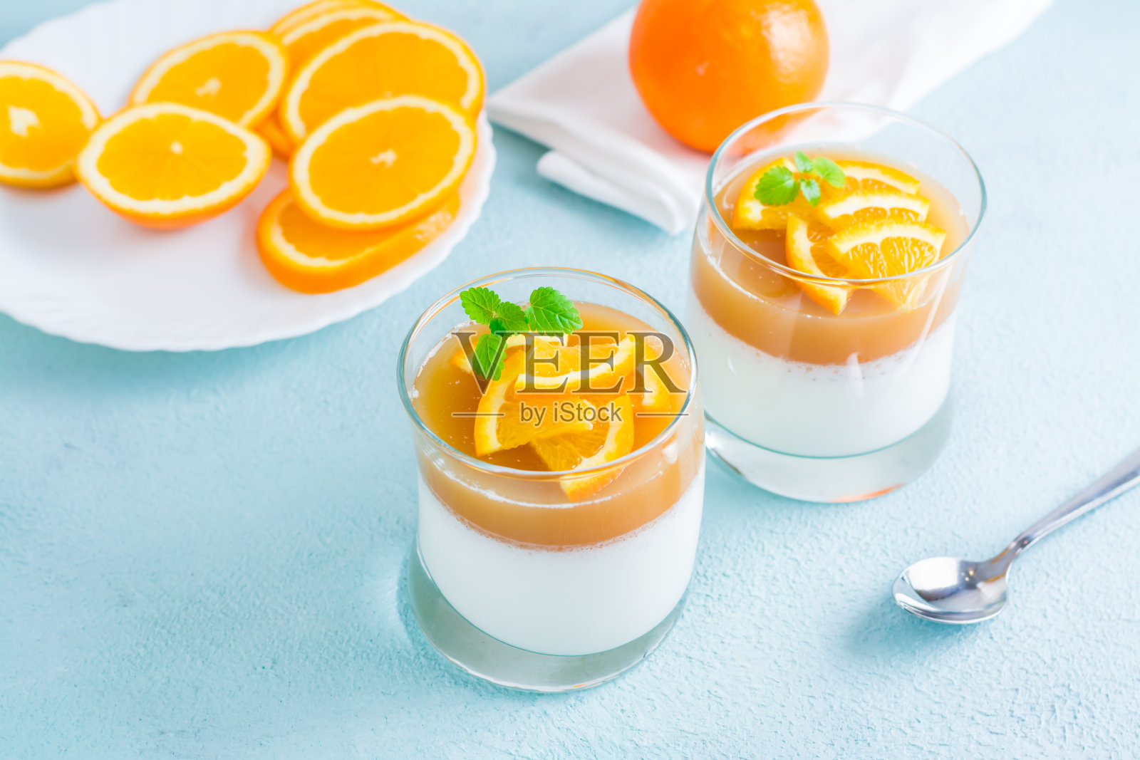 橙色意式奶冻,薄荷叶装在玻璃杯里,橙色切片放在浅桌上的盘子里。甜蜜的意大利甜点照片摄影图片