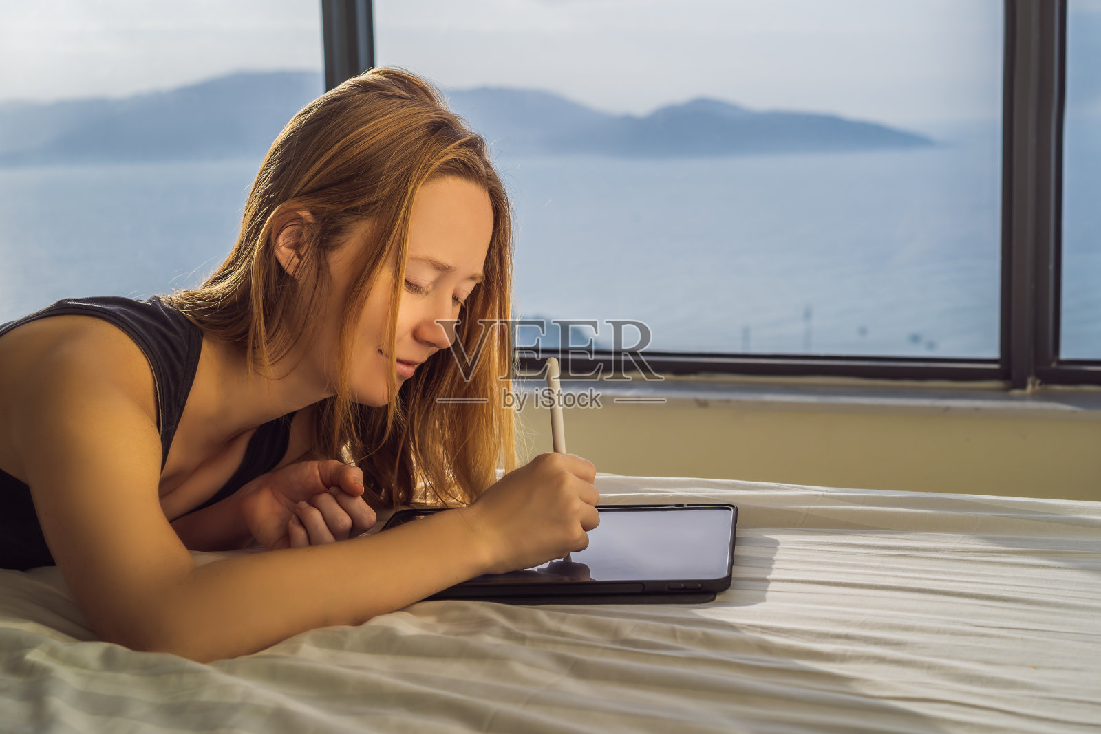 一名年轻女子在一扇海景窗的背景下画着平板电脑。设计师,素描照片摄影图片