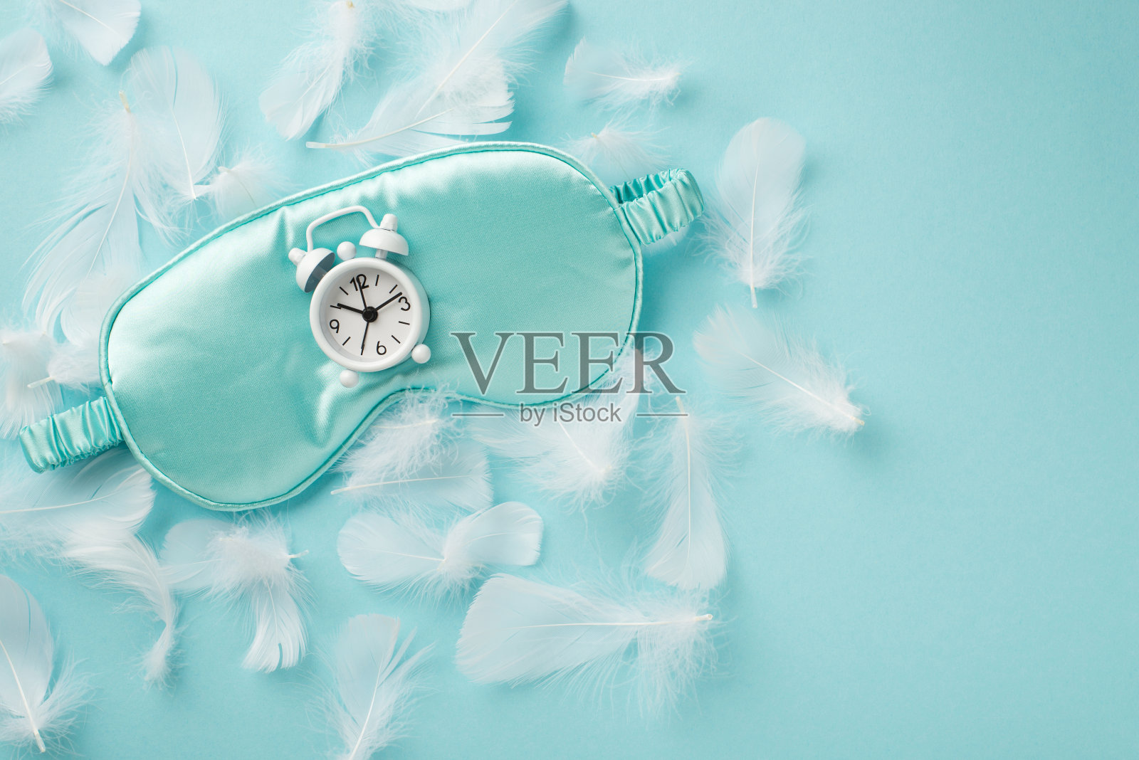 俯视图照片的蓝色丝绸眼罩与白色时钟上的许多白色嫩羽毛散落在柔和的天空蓝色孤立的背景空白空间照片摄影图片
