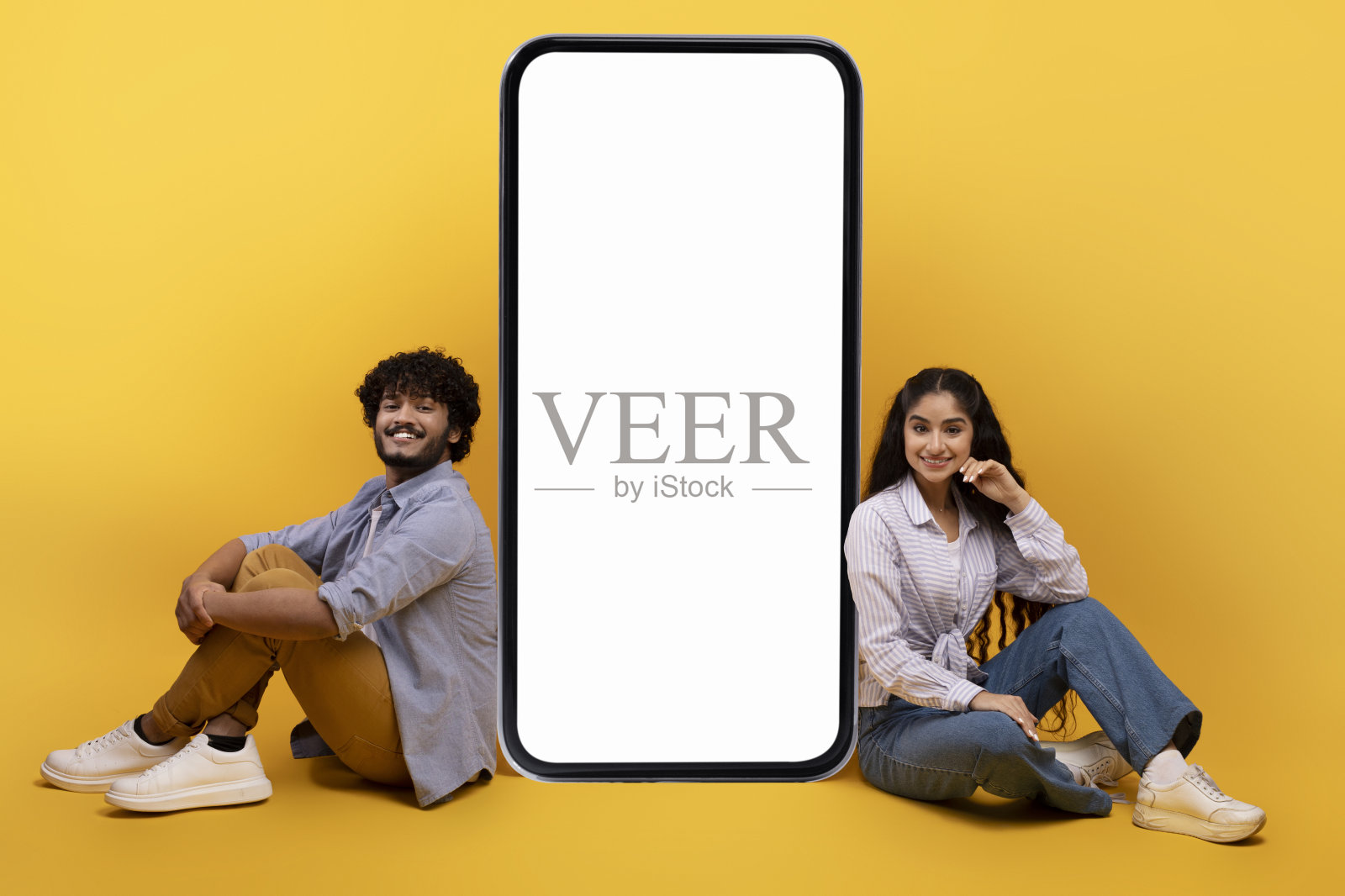 一对激动的印度夫妇坐在空白屏幕的巨型智能手机旁,展示你的广告复制空间照片摄影图片