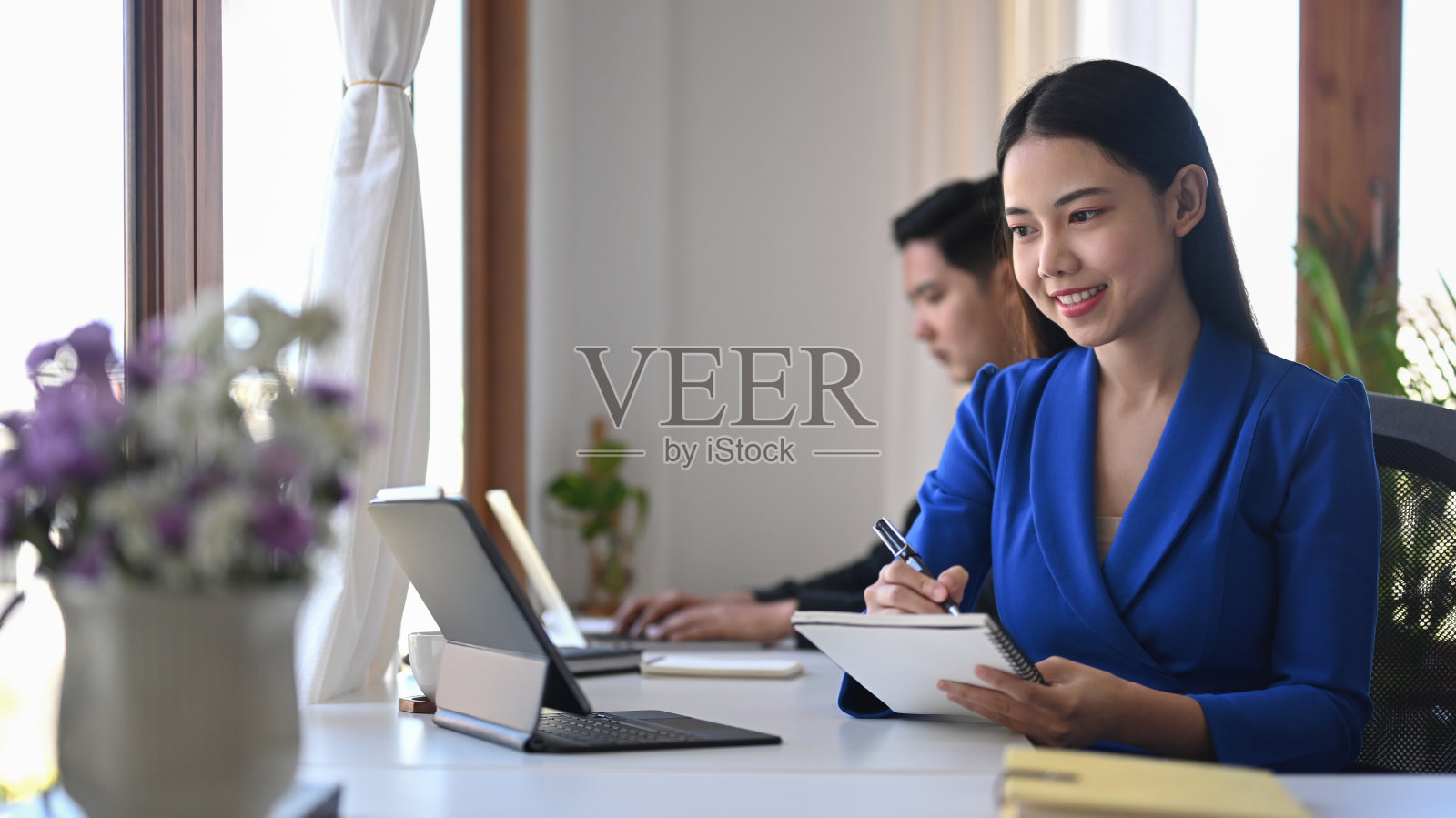 美丽的亚洲女人看着电脑平板电脑,在笔记本上做笔记。照片摄影图片