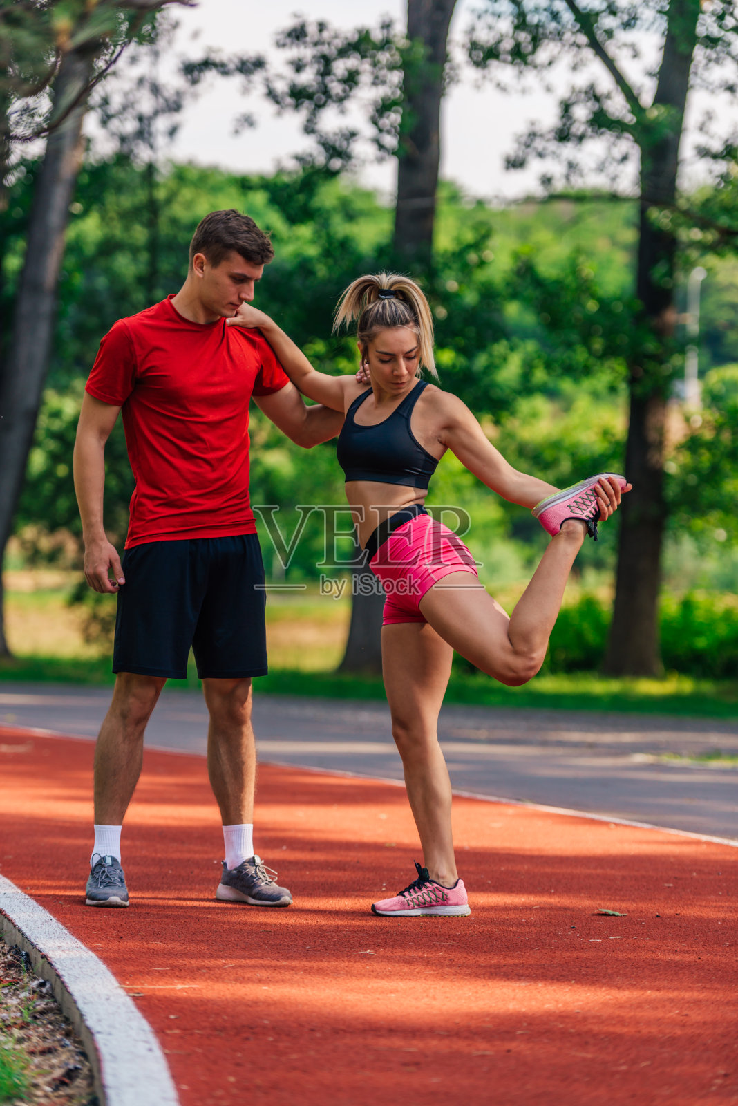 年轻的男运动员在他的年轻的女搭档在跑道上跑步后伸展腿时支持她。照片摄影图片