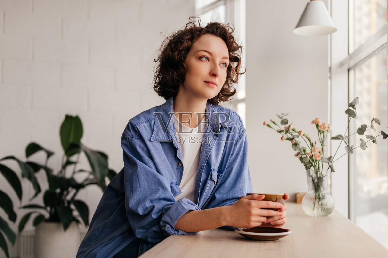 一头卷发的年轻女子微笑着坐在咖啡馆里喝着咖啡,望着室内的窗户照片摄影图片