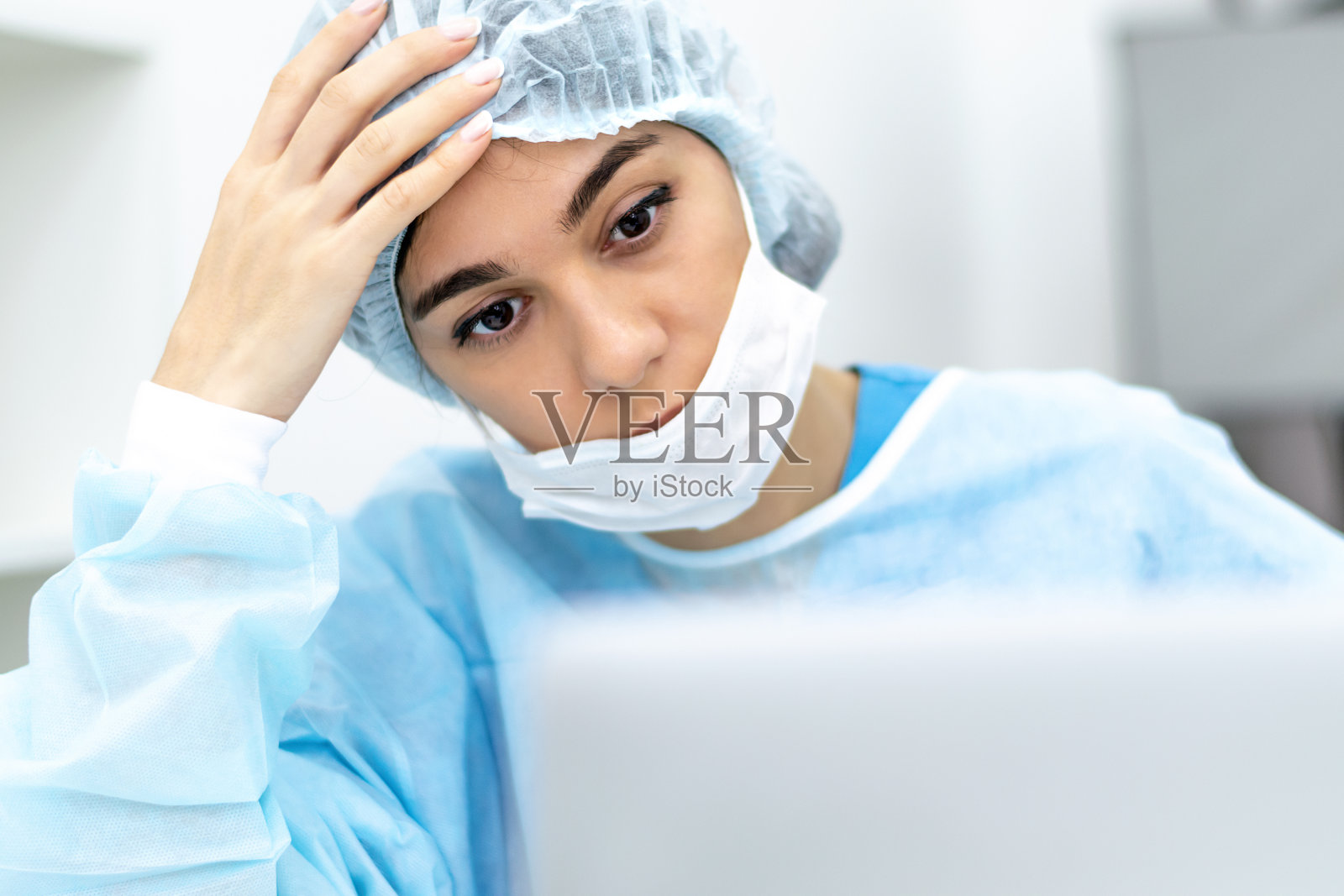 一位疲惫的女医生戴着口罩和手术服坐在医疗办公室的笔记本电脑屏幕前。医疗保健概念照片摄影图片
