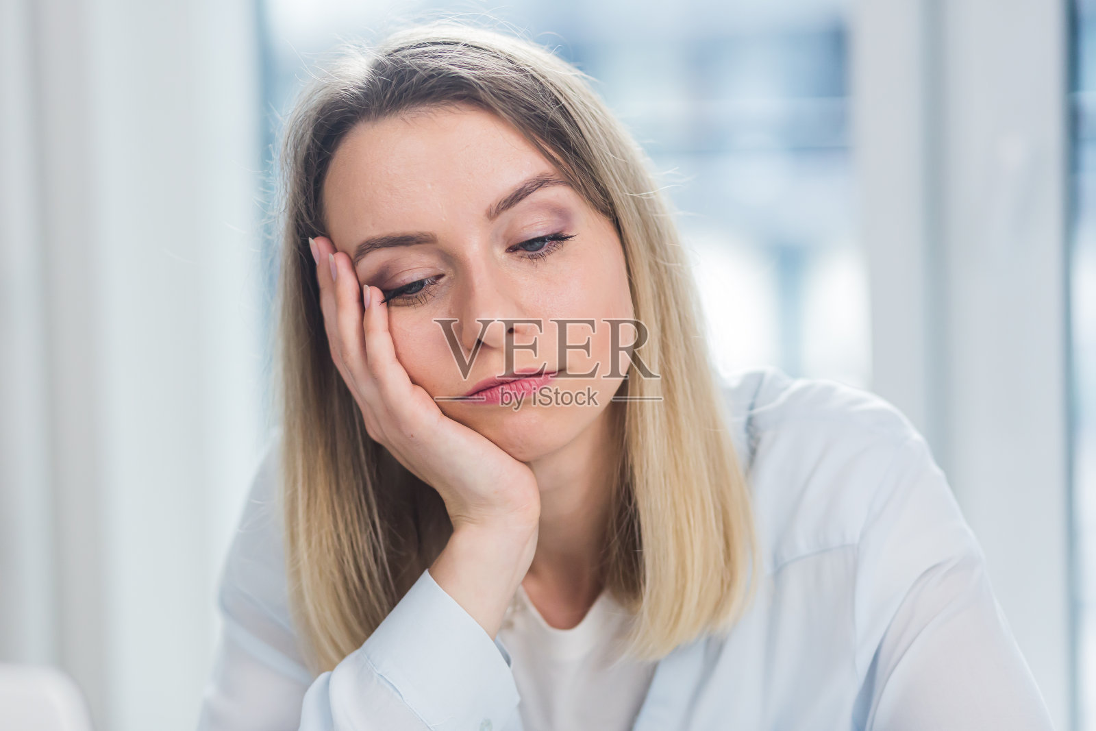 一个年轻的成年金发女商人的特写肖像看着在办公室或在蓝色窗户背景的室内的相机。在室内穿着正式服装面对自信的女经理照片摄影图片