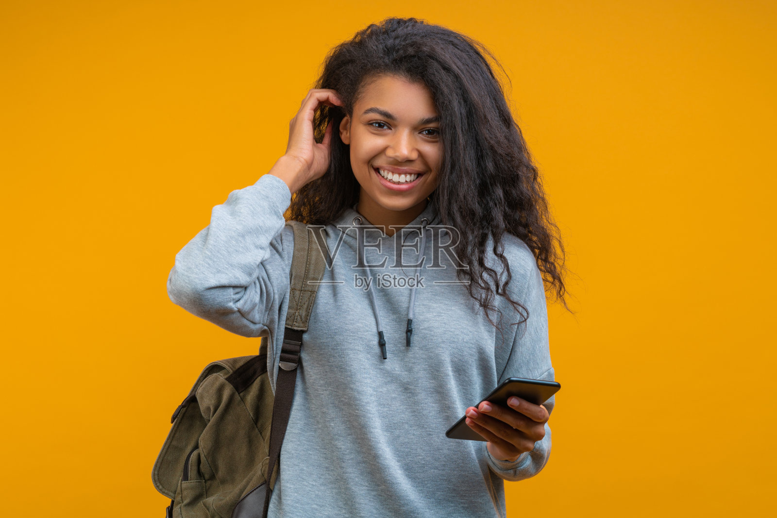 可爱的微笑,穿着随意的年轻学生女孩拿着手机在手里,固定她的头发,孤立在黄色的背景照片摄影图片