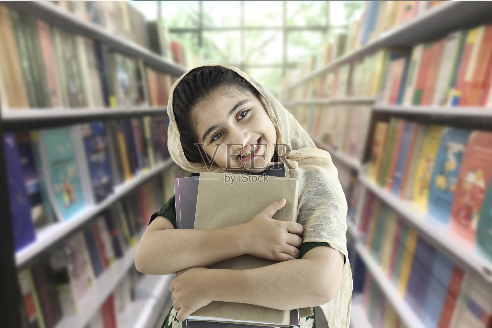 可爱的微笑巴基斯坦穆斯林学生女孩美丽的眼睛戴着头巾,站在图书馆或书店的书柜模糊的背景和许多书。照片摄影图片