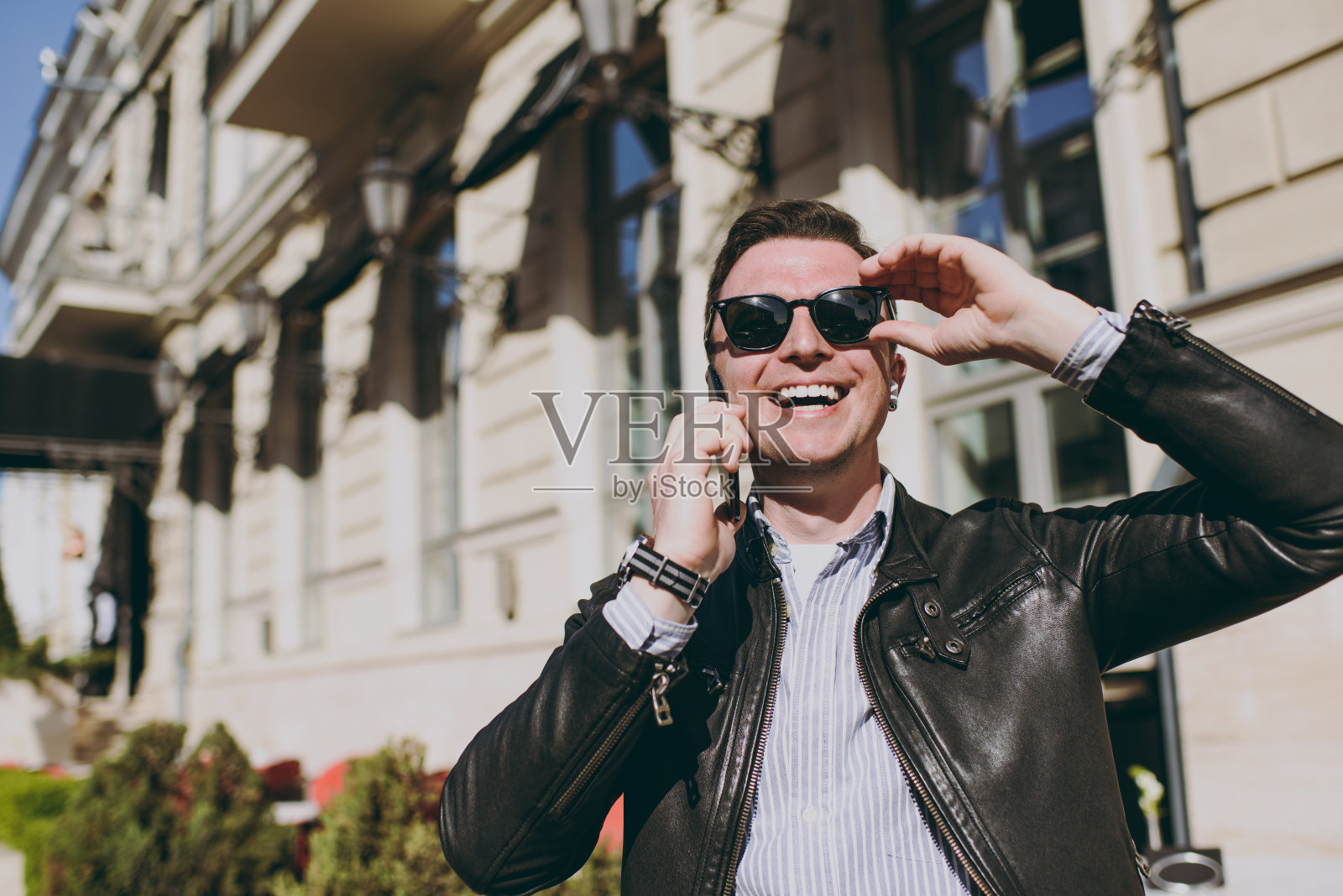 年轻微笑积极有趣白种人快乐男子20多岁穿皮夹克触摸眼镜说话用手机说话散步户外概念人都市生活春天。照片摄影图片