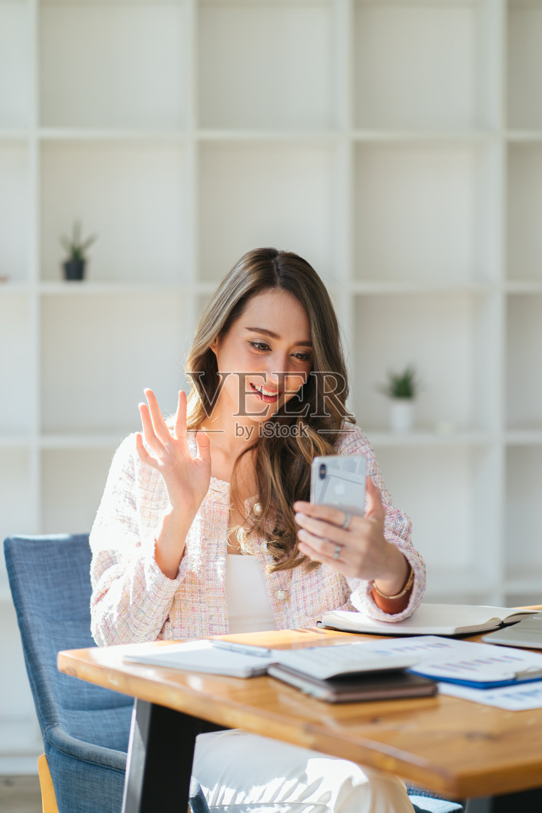 快乐的年轻商业亚洲妇女坐在木桌在家里拿着手机看着屏幕挥动的手视频呼叫远程朋友在线在移动聊天应用使用智能手机视频聊天应用程序。照片摄影图片