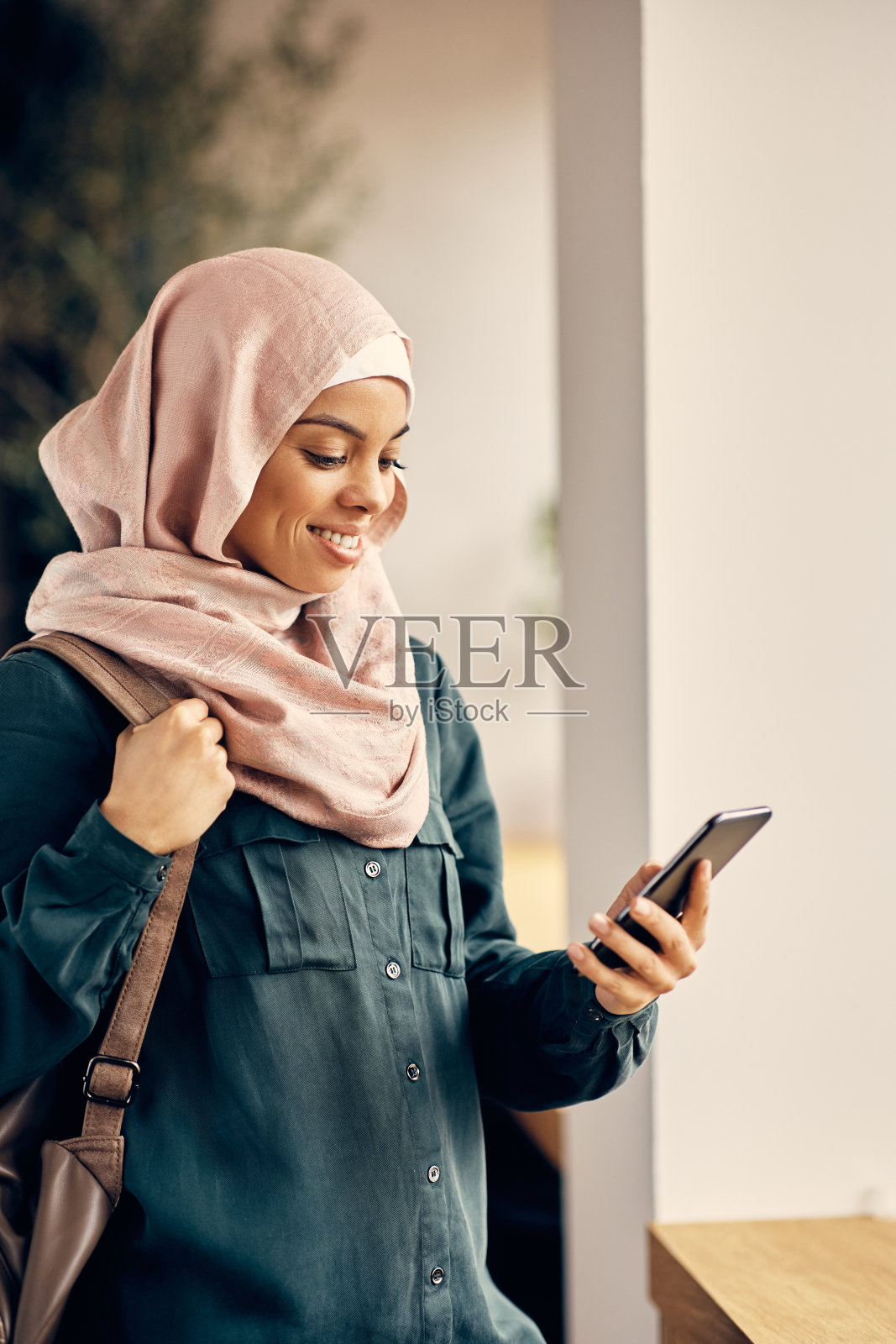 快乐的女学生戴着头巾,在大学的走廊里用手机发短信。照片摄影图片