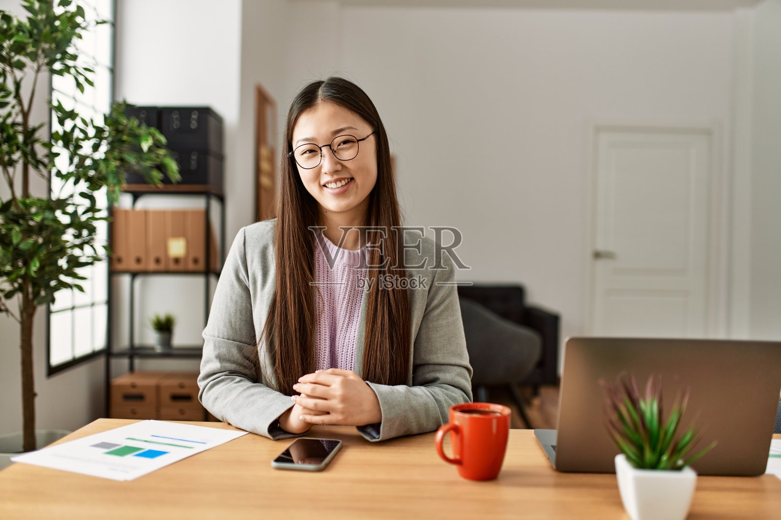 年轻的中国商务人士穿着商务风格的衣服,坐在办公室的办公桌上,看起来积极和快乐,站着微笑,自信的微笑,露出牙齿照片摄影图片