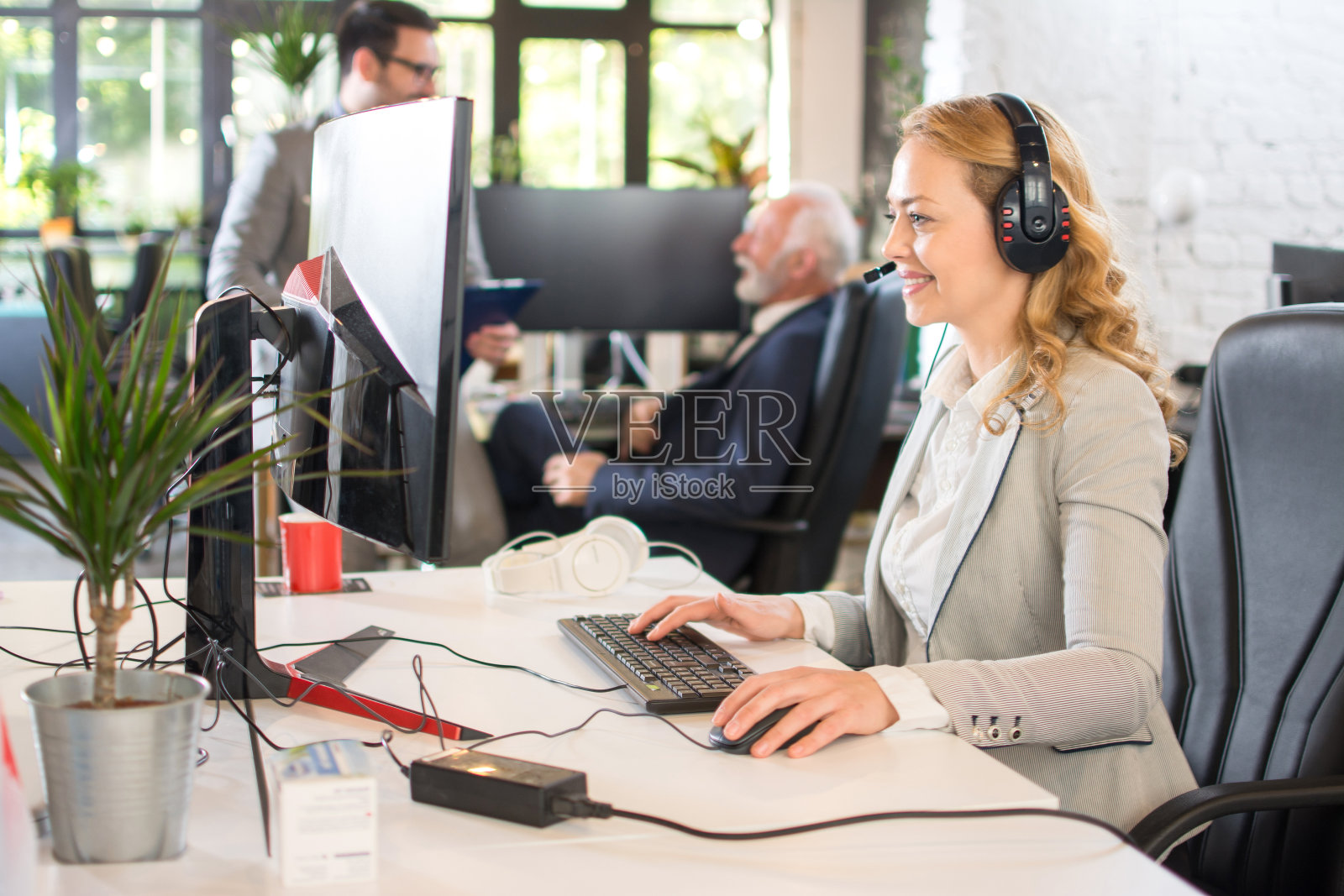 快乐成熟的女商人戴着耳机,看着办公室里的电脑在电话会议上说话照片摄影图片