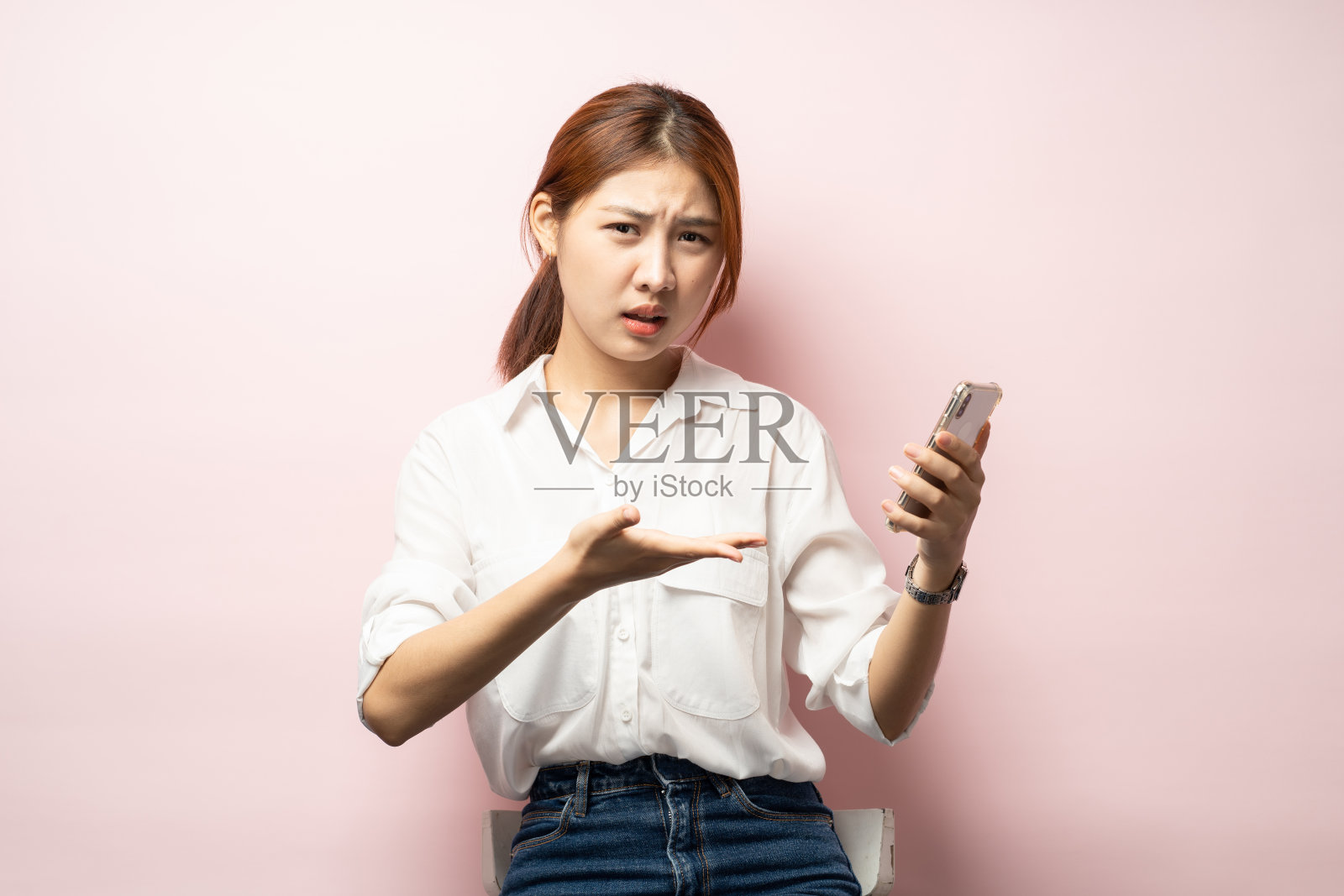 沮丧的亚洲妇女抱怨智能手机,拿着手机,看起来困惑,粉红色的背景。照片摄影图片