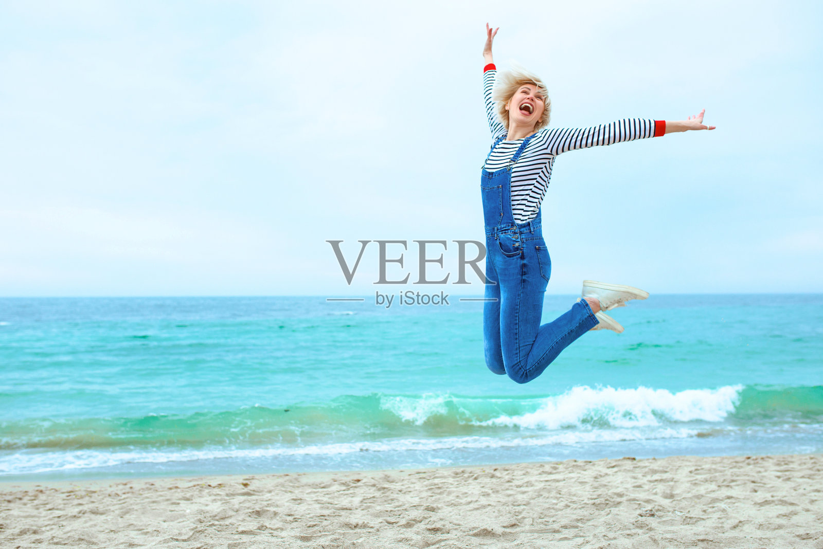 美丽的年轻金发白人妇女在度假条纹衬衫,运动鞋和牛仔布整体跳在惊人的蓝色海洋背景的海滩上照片摄影图片