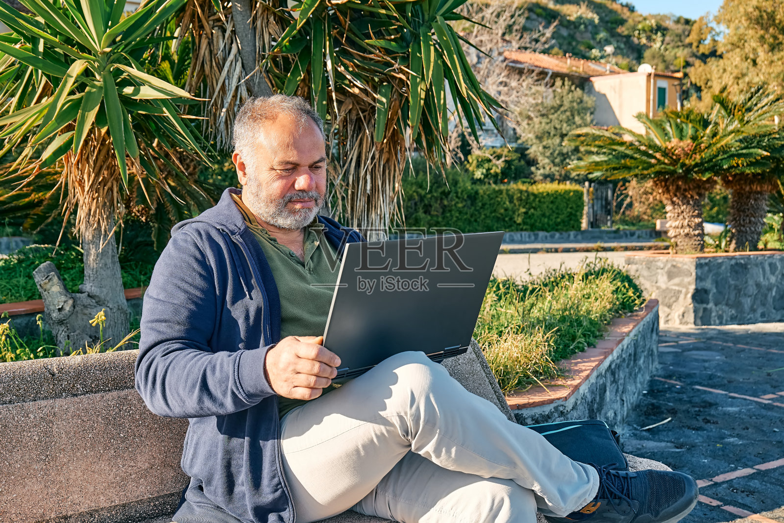 英俊的大胡子成熟男人,坐在海边的长椅上,带着笔记本电脑在户外工作。现代生活方式,联系,商业,自由职业的概念。照片摄影图片