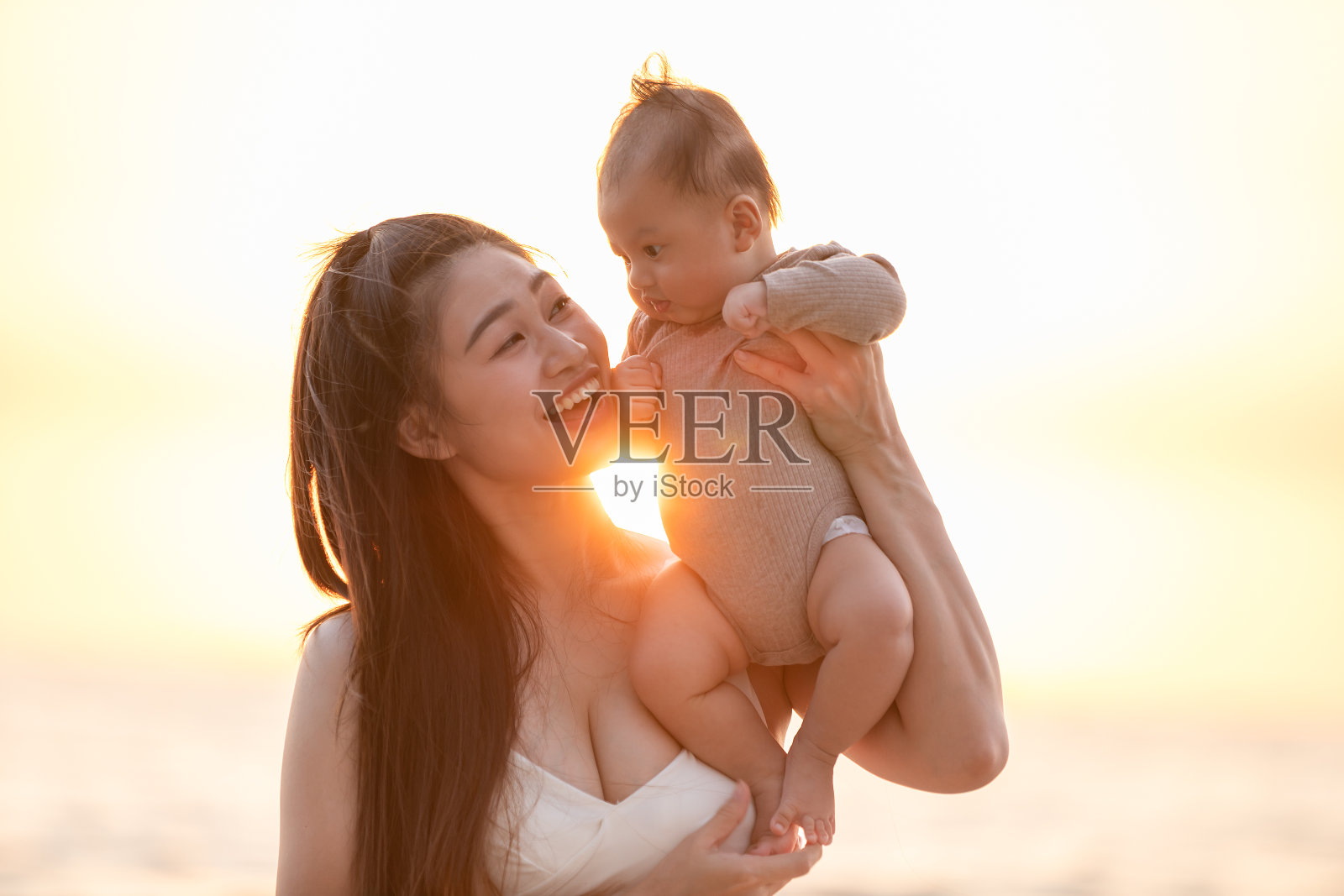 美丽迷人的亚洲妈妈抱着她的新生儿在手和吻在婴儿的头甜美可爱的户外海滩上。快乐的妈妈和小宝宝一起看着夕阳自然的微笑照片摄影图片