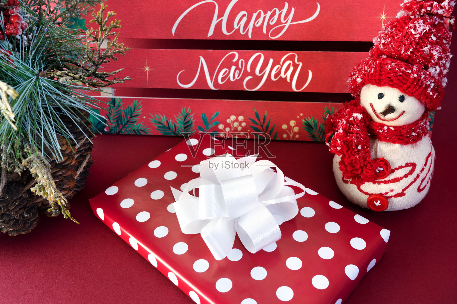 写着“新年快乐”的新年贺卡,一个带着白色蝴蝶结的礼盒,一个戴着红色帽子和围巾的雪人,红色背景上的松树、球果和红色浆果构成的冬季图画照片摄影图片