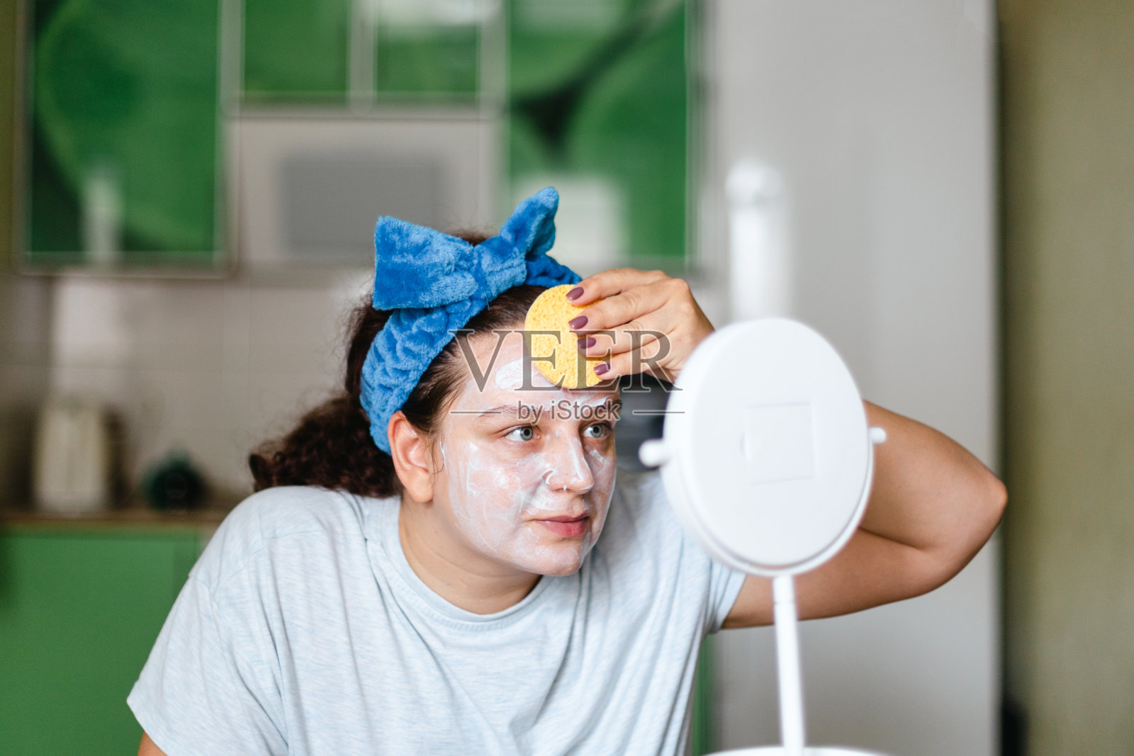 白种人妇女从脸上洗去保湿面膜,头上戴着蓝色蝴蝶结,看着镜子。谈论照顾自己和皮肤。青春延伸——抗衰老。家庭美容化妆程序照片摄影图片