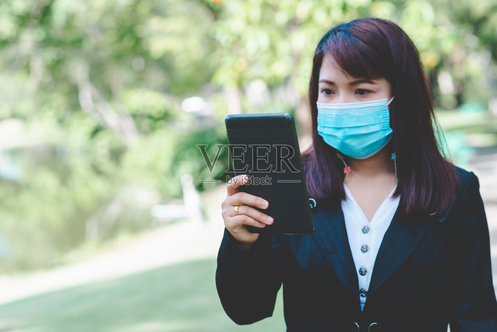 女商人戴着口罩,用智能手机平板电脑在屏幕上打字,在公园里用应用程序设备工作,伴着日出和模糊的自然背景。冠状病毒或covid-19的业务新常态。照片摄影图片