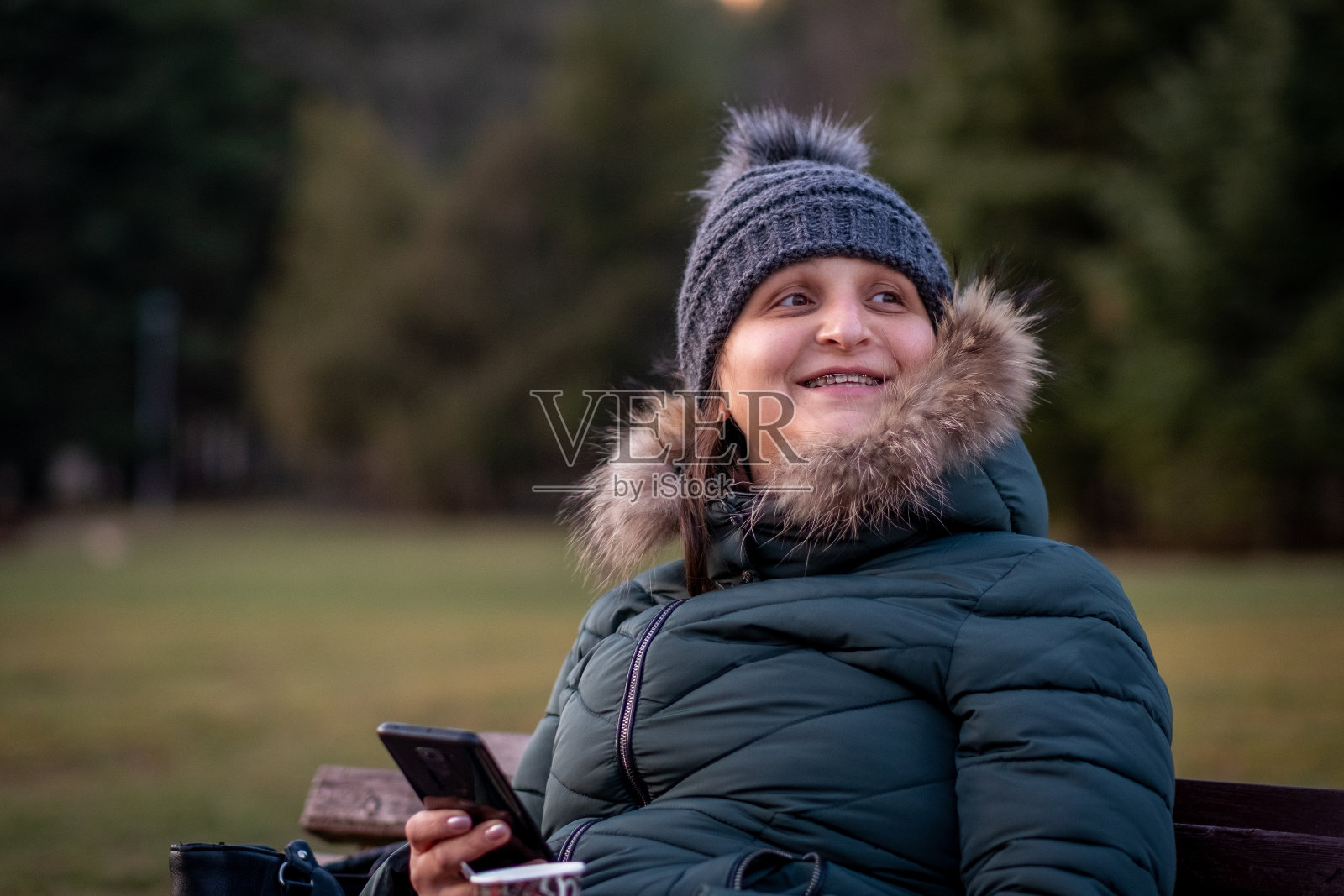 一个穿着夹克,戴着帽子,戴着蓬松的兜帽,喝着咖啡,拿着手机的快乐的女孩看着坐在公园长椅上的朋友们照片摄影图片