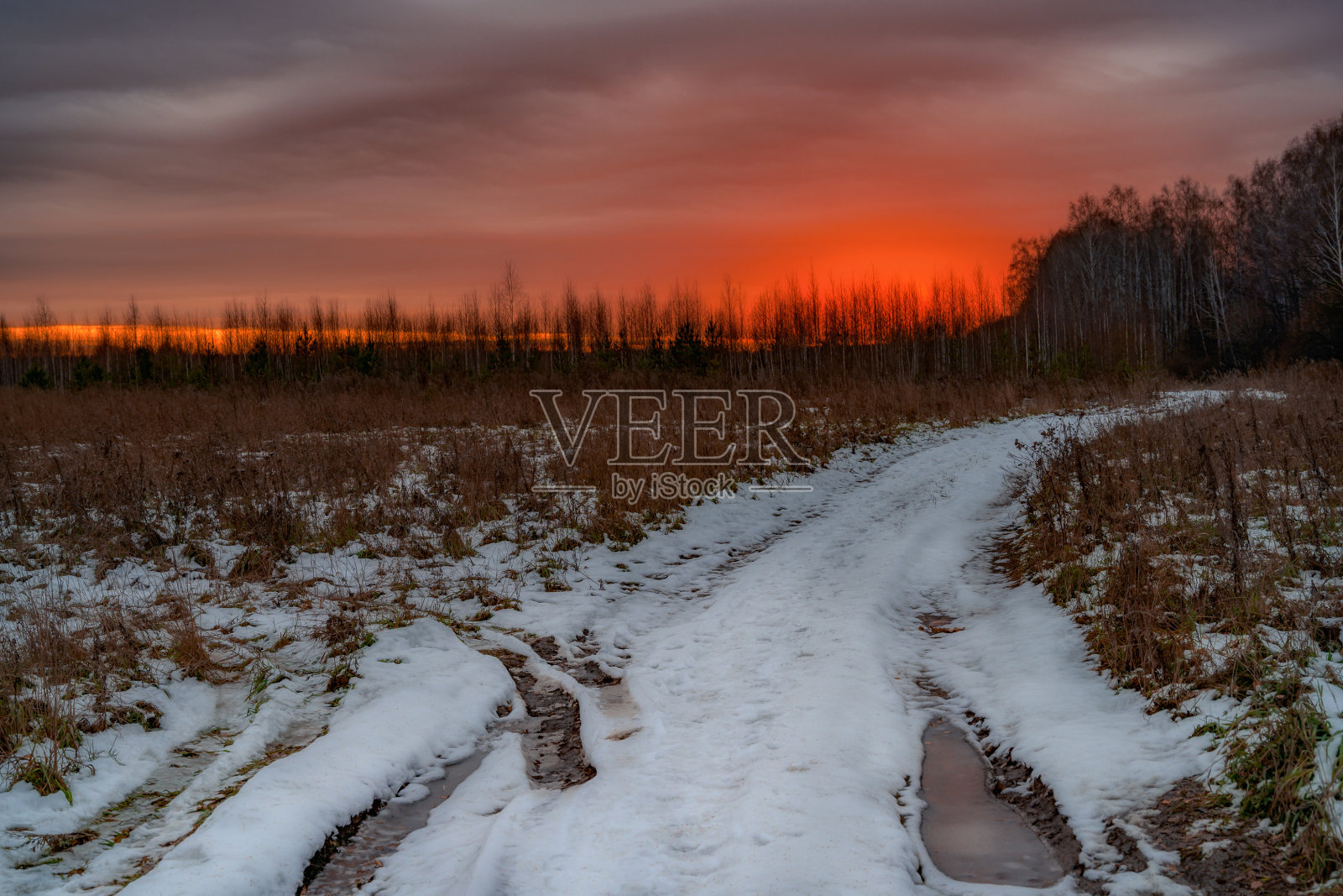 一条乡村小路,在秋天解冻时还没有完全冻住。红色调的天空,压着雨夹雪,水坑,一片森林照片摄影图片