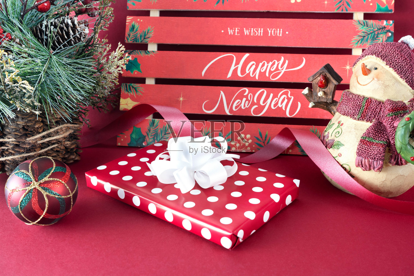 圣诞背景:一个带蝴蝶结的礼盒,一个戴着帽子和围巾的雪人,一个圣诞树玩具,一个冬天用松果装饰的圣诞树。红色背景的白色文字“新年快乐”照片摄影图片