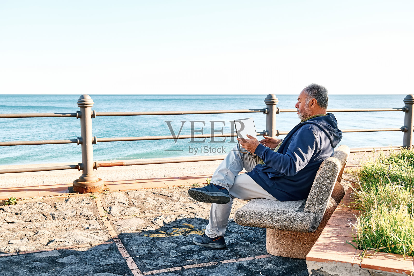 英俊的大胡子成熟男人,坐在海边的长椅上,带着笔记本电脑在户外工作。现代生活方式,联系,商业,自由职业的概念。照片摄影图片