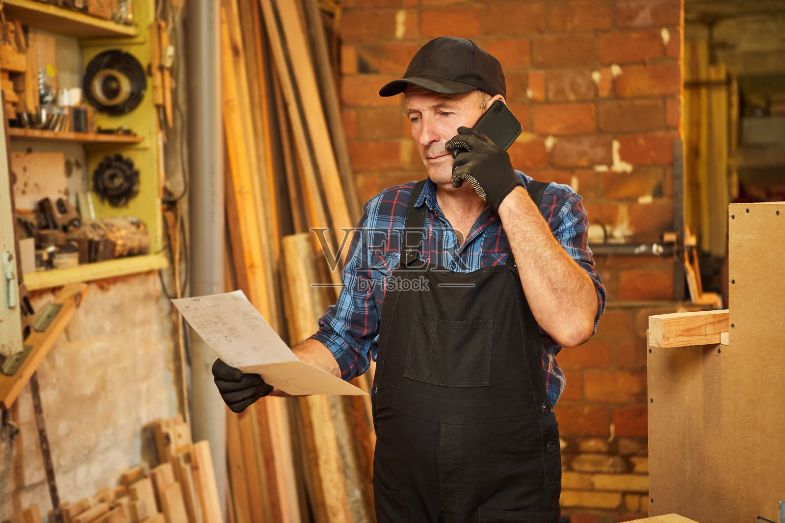 一名老年木匠的肖像,看着蓝图计划做一件家具,并与客户在木工车间的电话交谈照片摄影图片