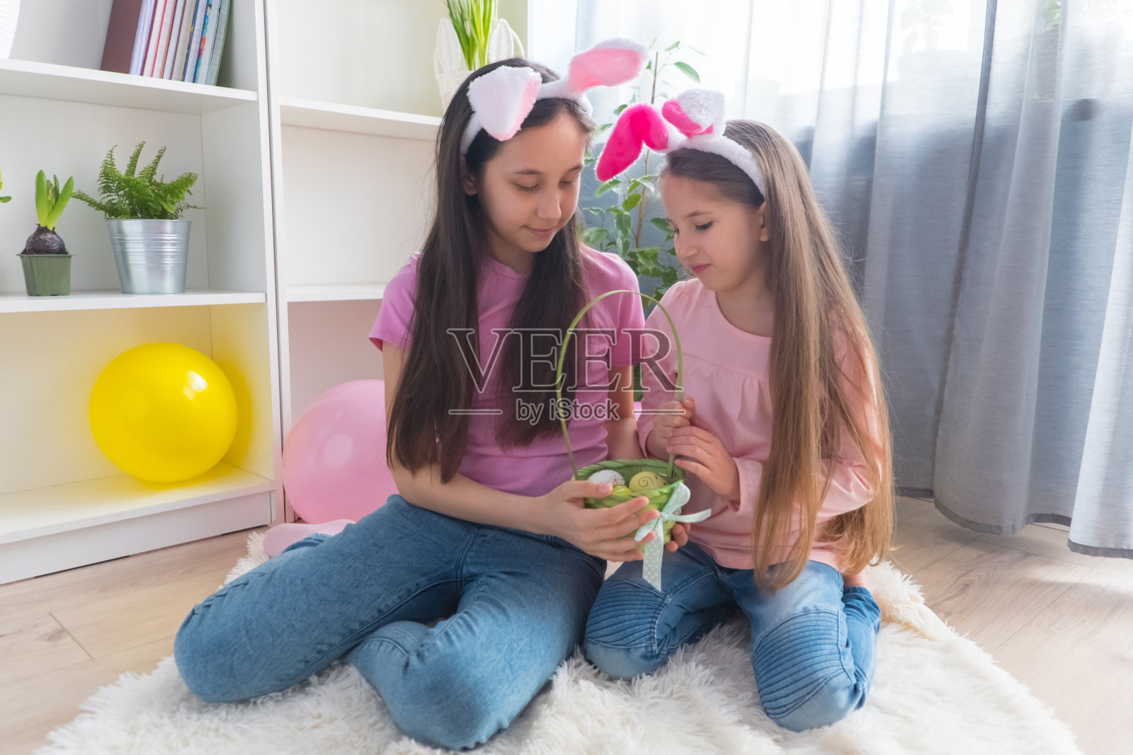 穿着粉红色t恤、耳朵戴着兔子耳朵、面带微笑的姐妹女孩在用气球装饰的房间里收集复活节彩蛋照片摄影图片